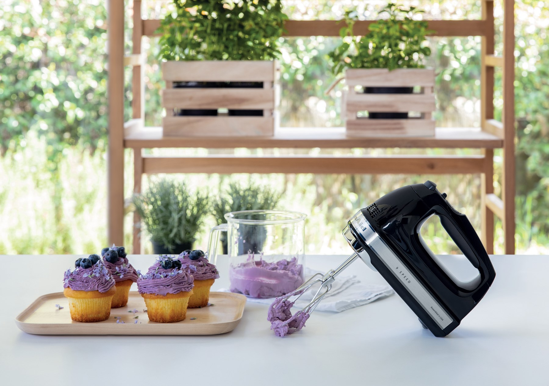 Una batidora de mano negra con varillas se sienta junto a cupcakes con glaseado púrpura y arándanos.