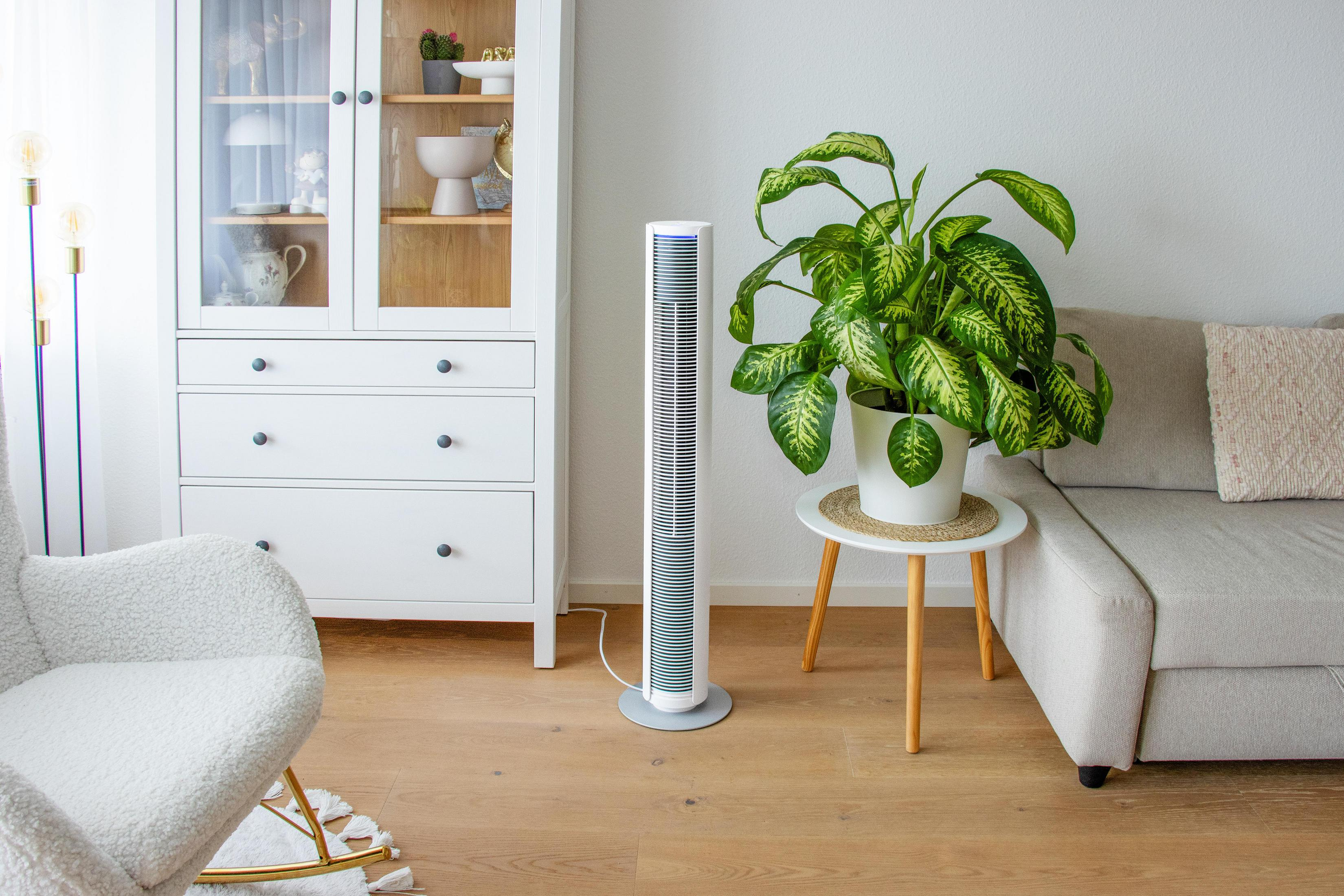 Un ventilateur fin et haut se tient près d'un canapé et d'une armoire.