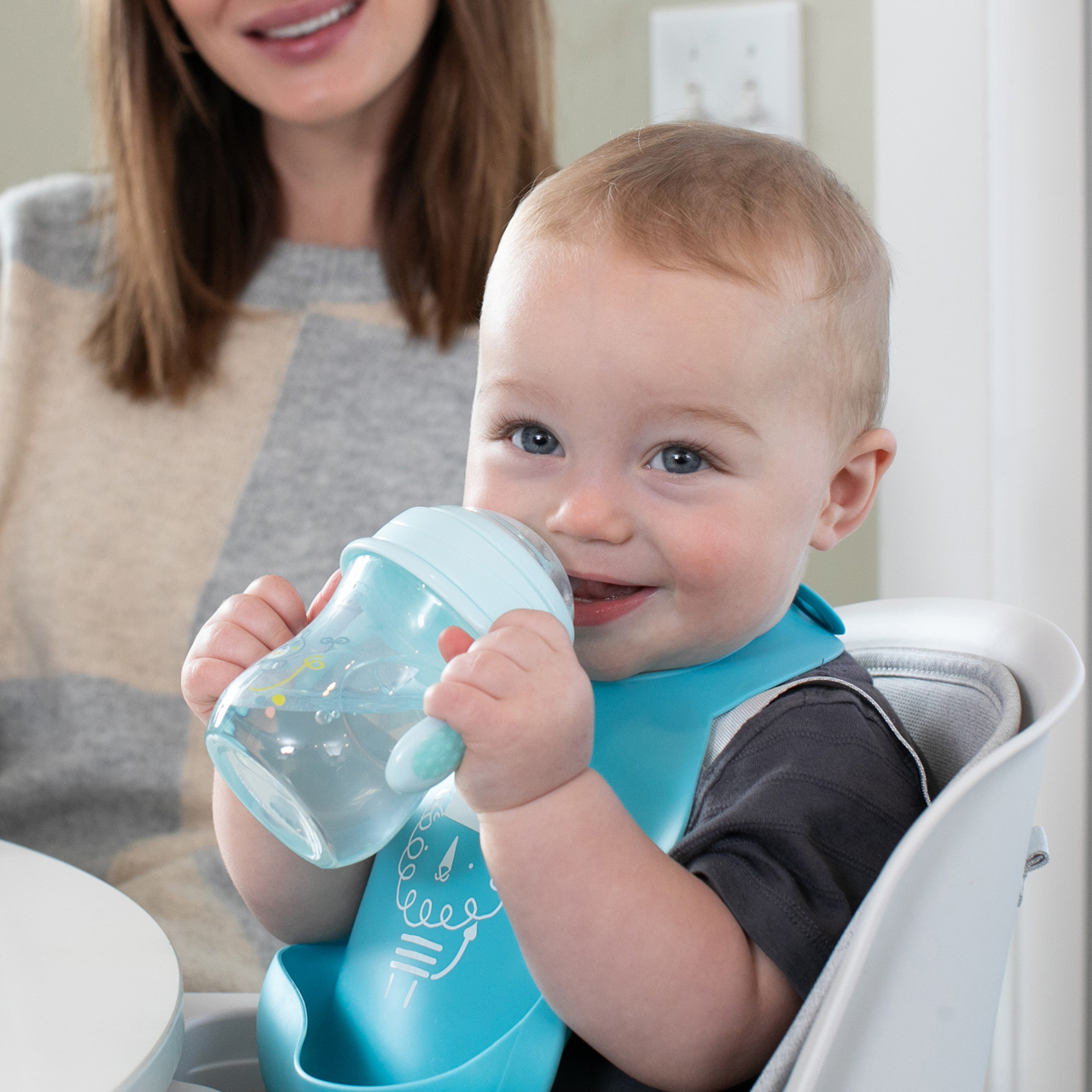 Ein Baby trinkt aus einer blauen Flasche, sitzt in einem Hochstuhl, trägt einen Latz.