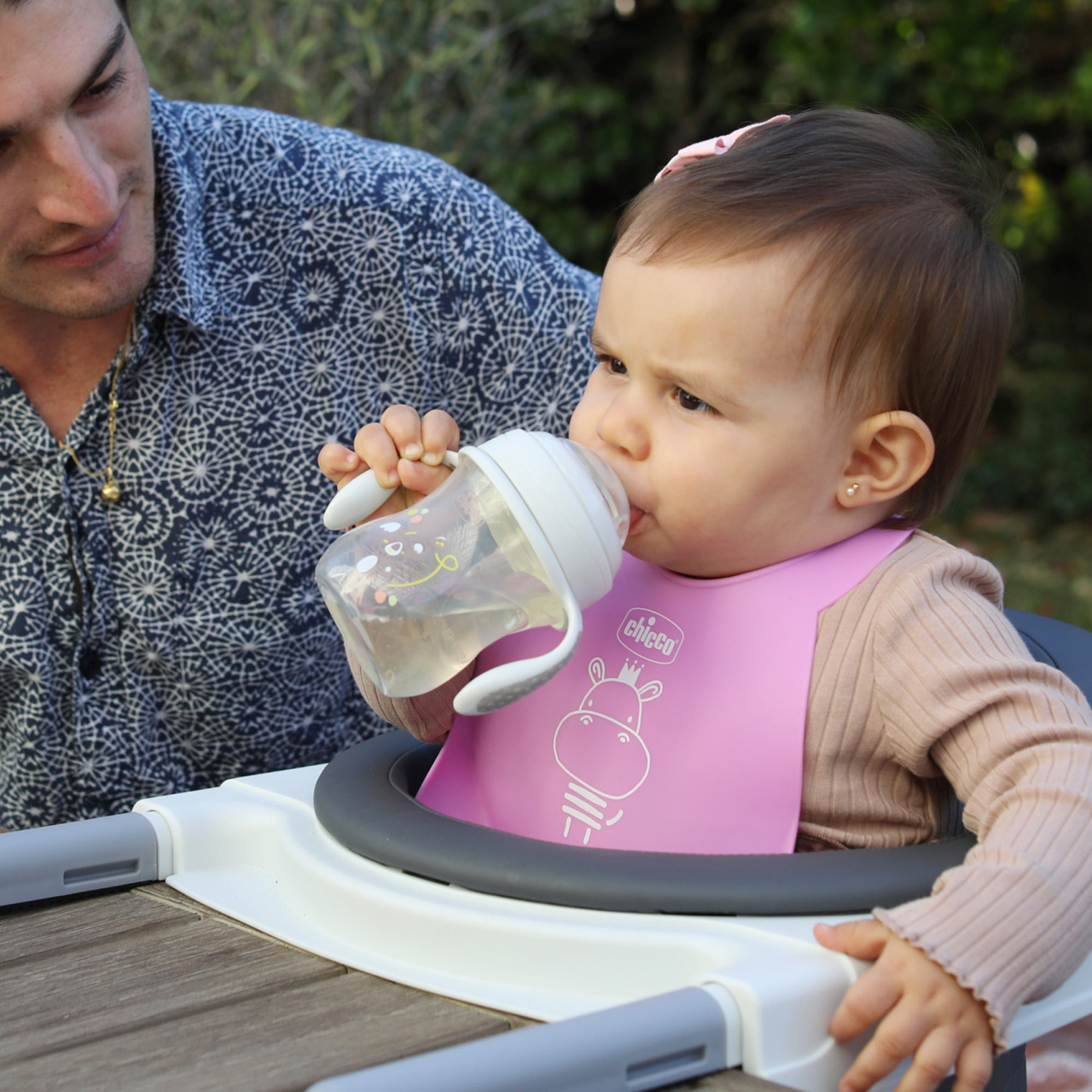 Ein Baby in einem Hochstuhl mit rosa Lätzchen trinkt aus einem durchsichtigen Schnabelbecher, ein Mann in einem gemusterten Hemd schaut ihr zu.