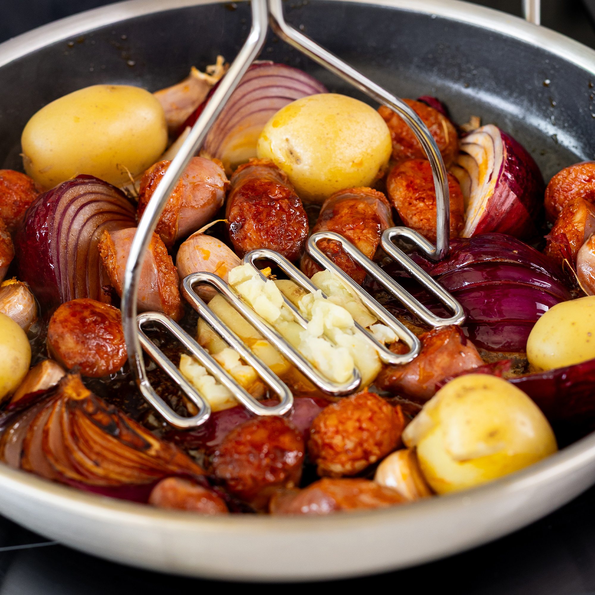 Bratwurst, Kartoffeln und rote Zwiebeln werden in einer Pfanne zerdrückt.