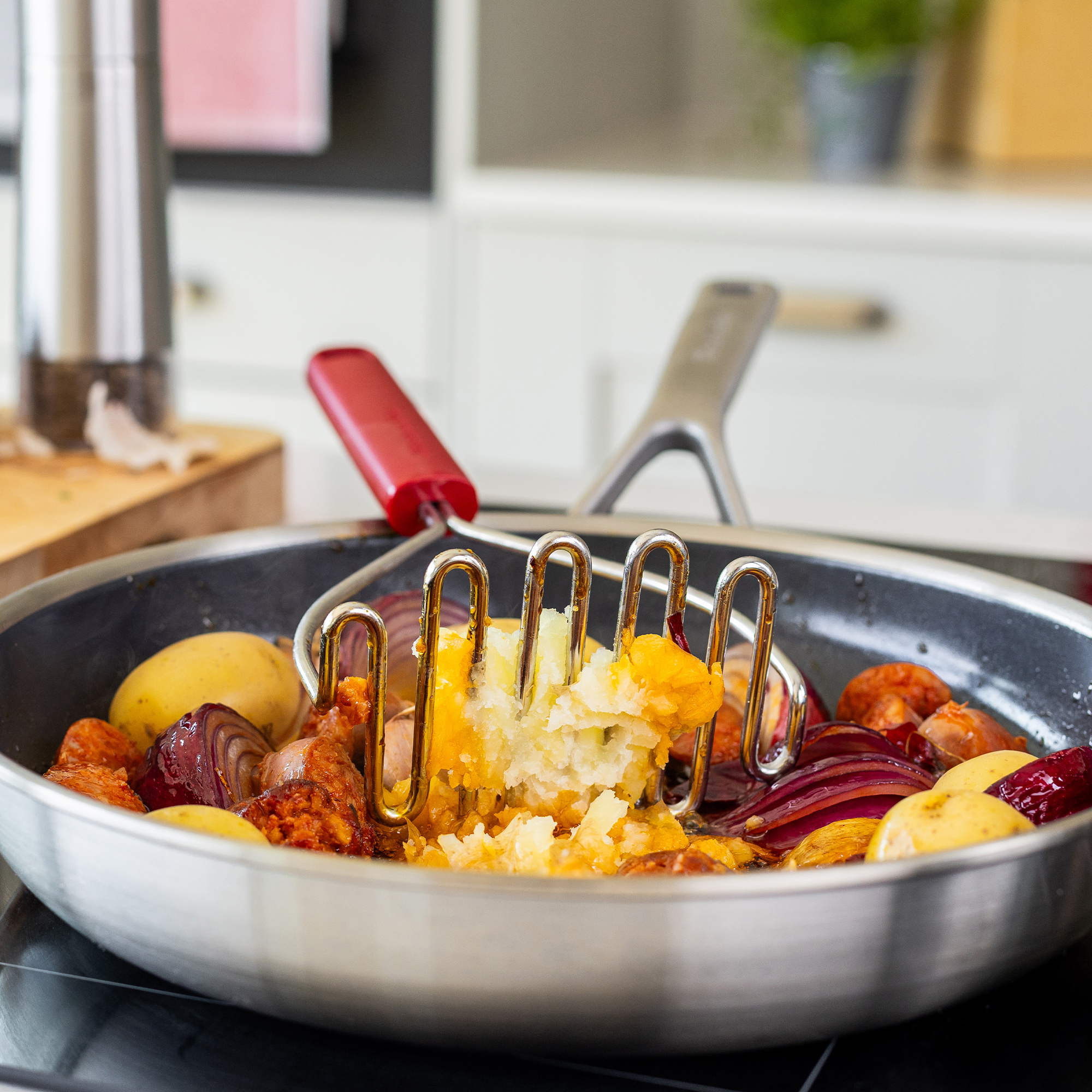 Ein Kartoffelstampfer zerdrückt Kartoffeln, Zwiebeln und Chorizo in einer Pfanne.