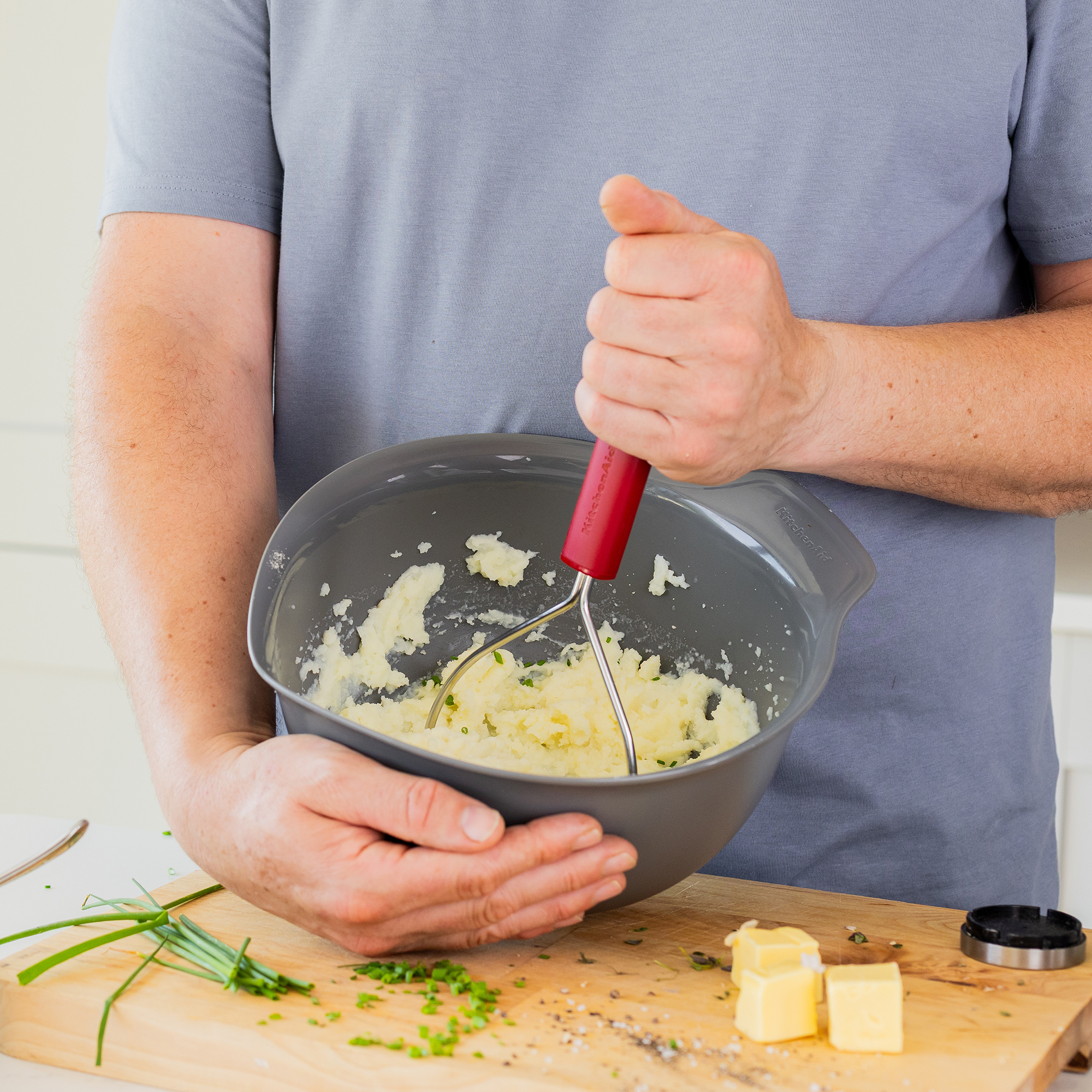 Eine Person zerdrückt Kartoffeln in einer grauen Schüssel mit einem Kartoffelstampfer.
