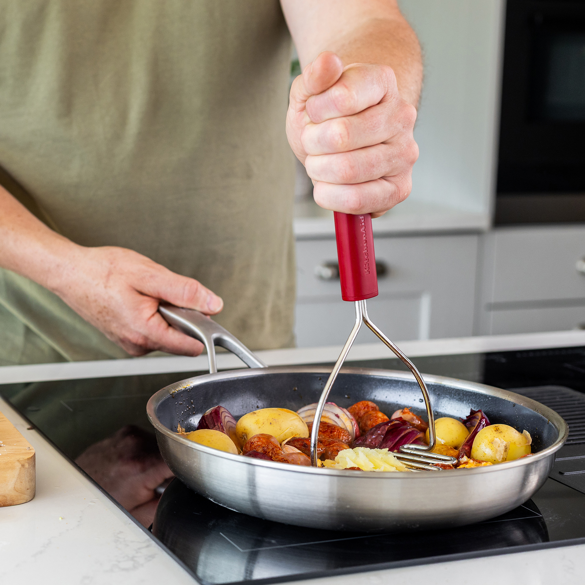 Eine Hand zerdrückt Kartoffeln in einer Pfanne mit Zwiebeln und Wurst.