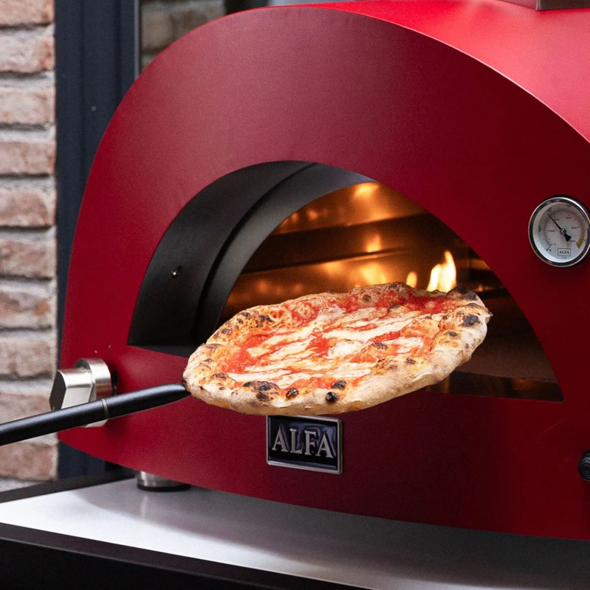 Eine Pizza wird aus einem roten Pizzaofen genommen. Sie hat Tomatensoße und Käse.