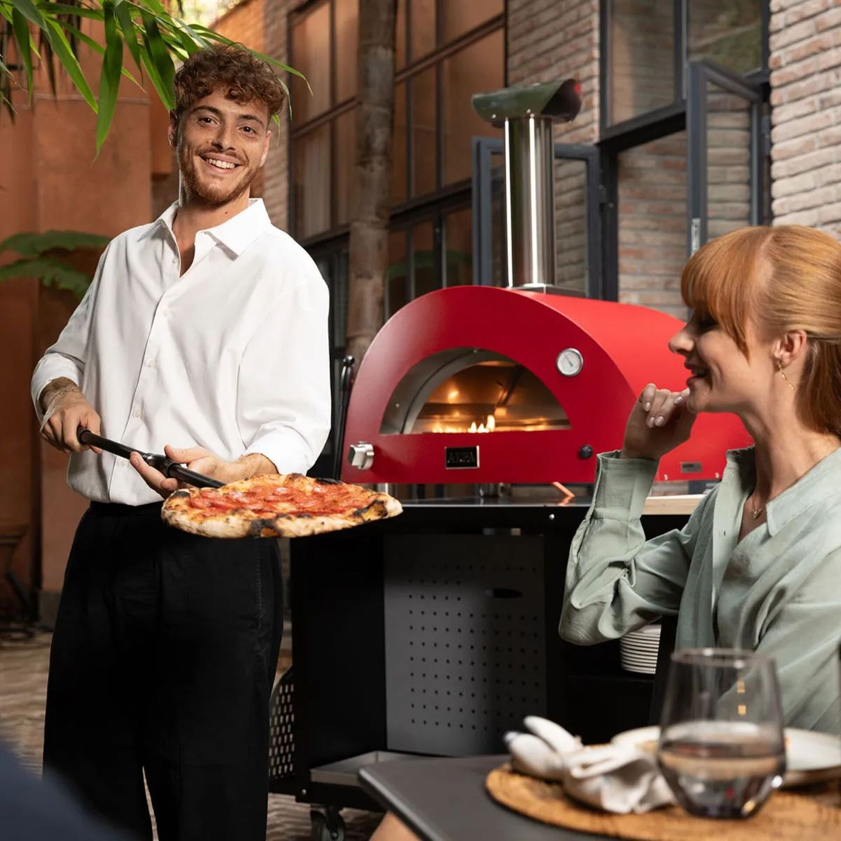 Mann mit Pizza vor rotem Ofen, Frau sitzt am Tisch.