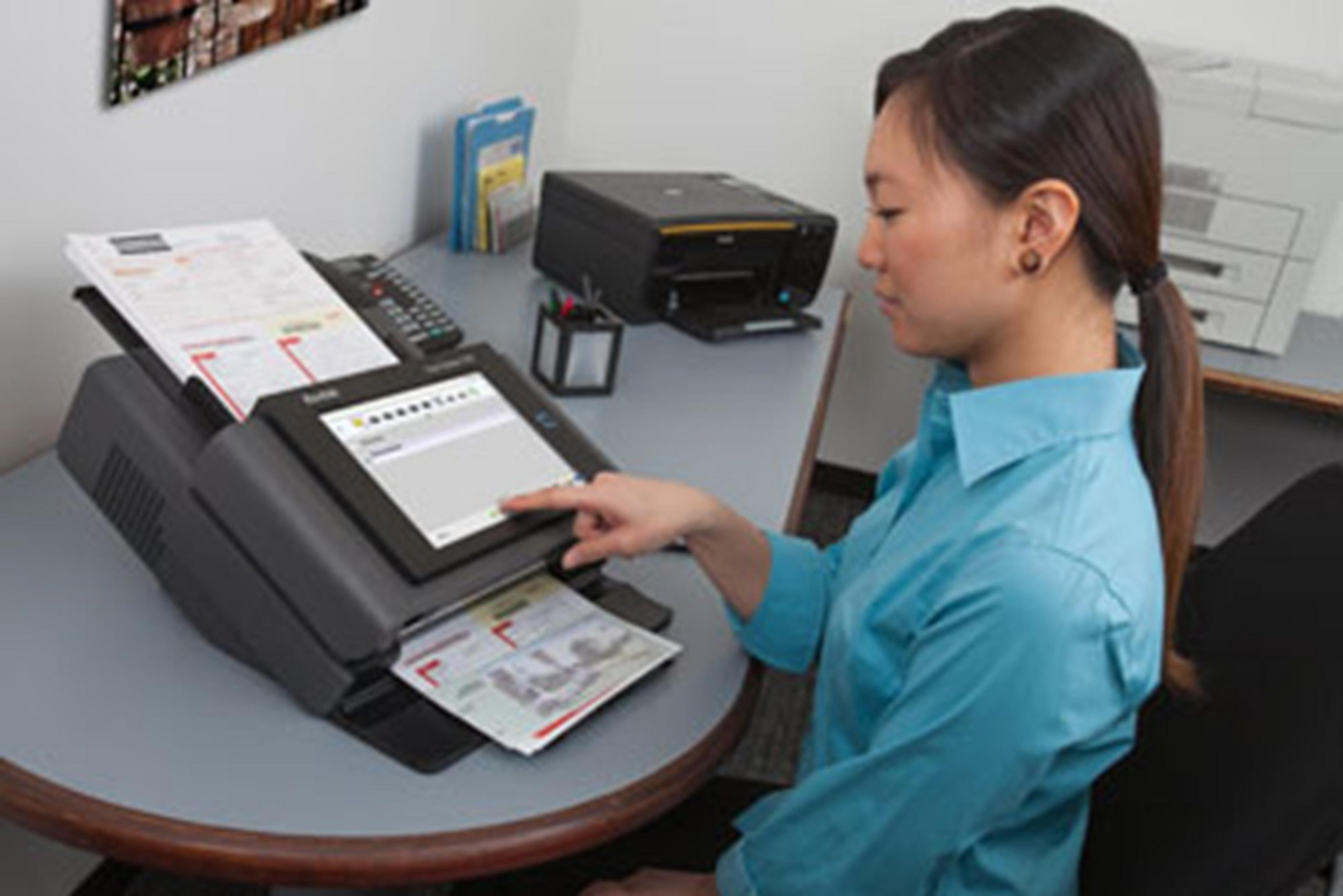 Kobieta używa skanera przy biurku, obok jest drukarka i telefon.