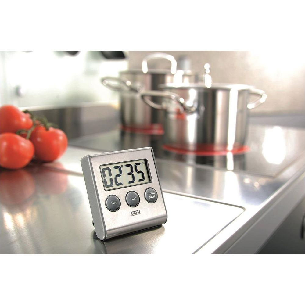 Ein digitaler Timer steht auf einer Metalloberfläche neben Tomaten. Kochtöpfe sind im Hintergrund.