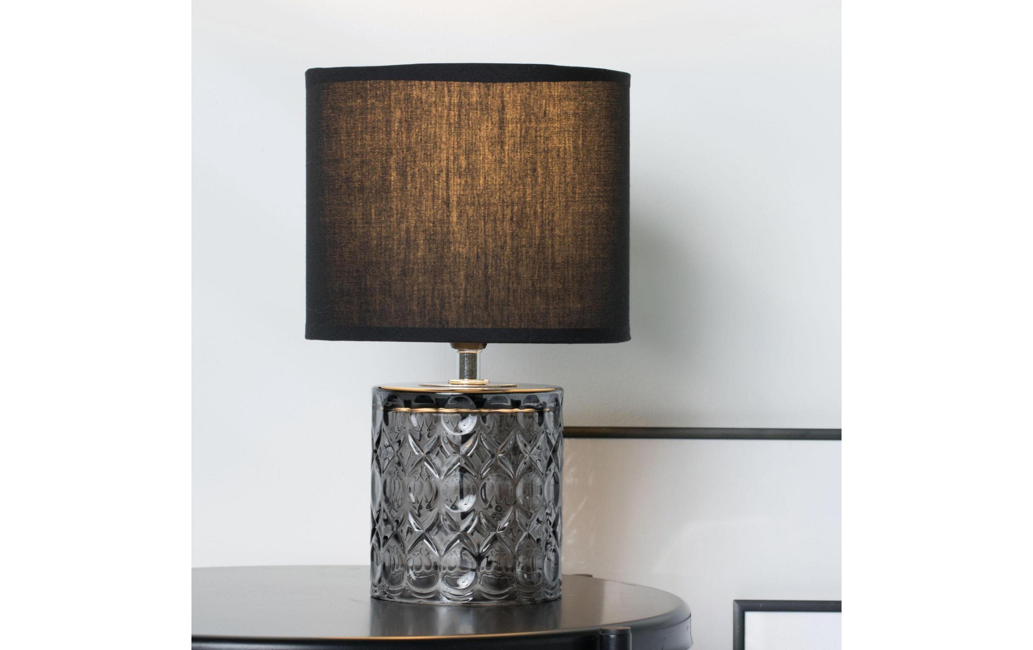 Szara lampa z czarnym kloszem na ciemnym stole przy białej ścianie.