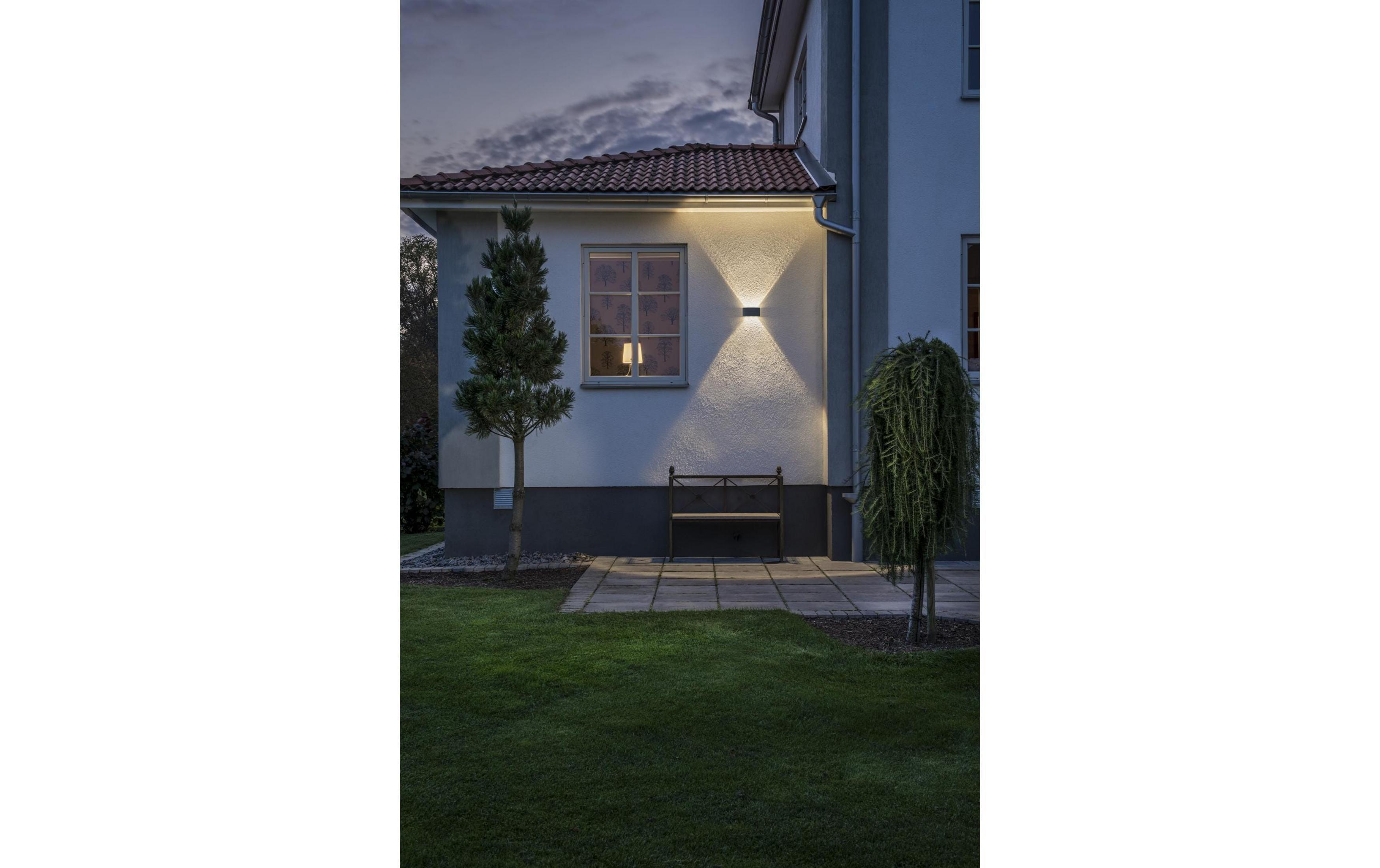 Esterno di una casa al crepuscolo, con finestra illuminata, panchina e alberi.