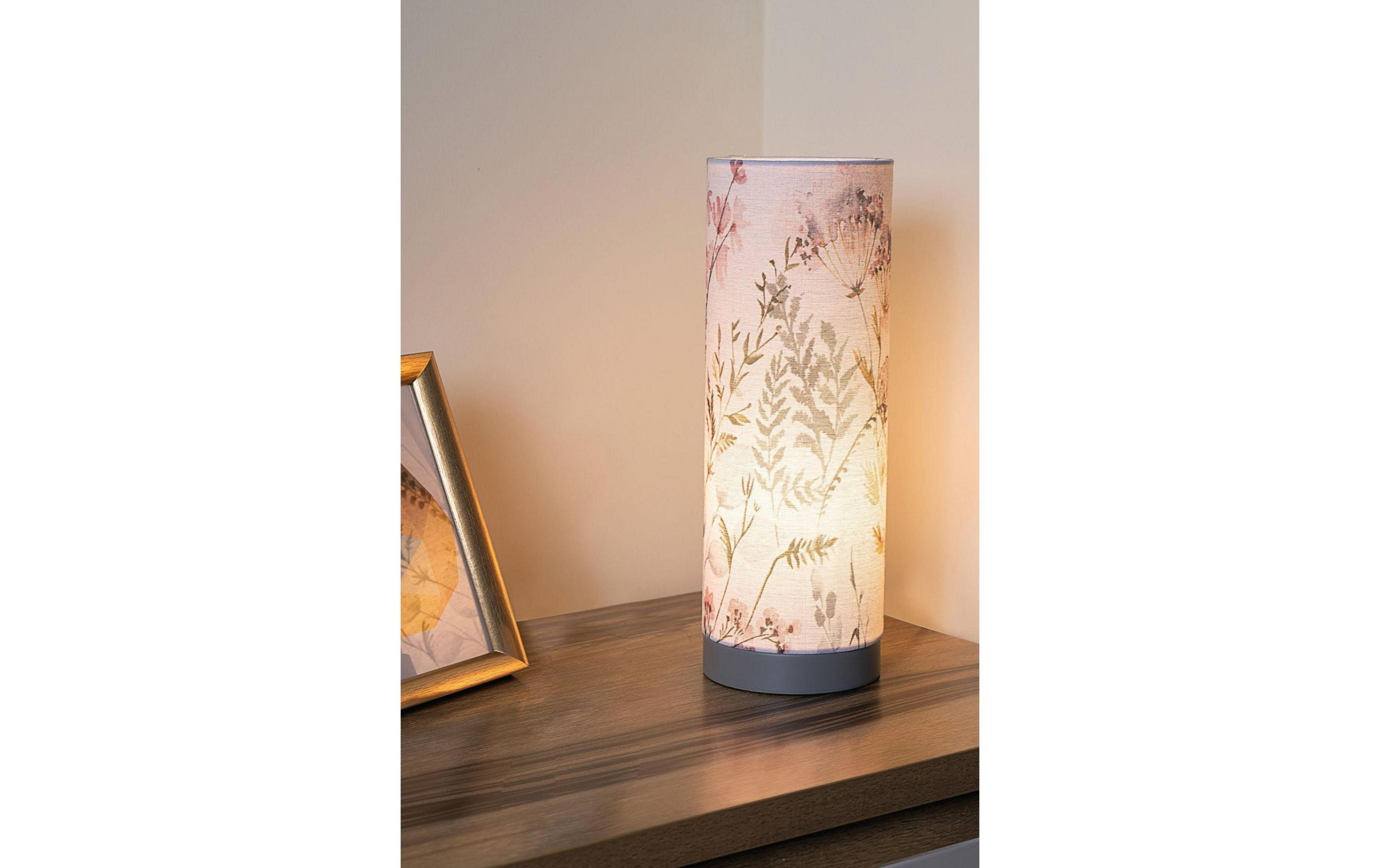 Cylindryczna lampa z motywem kwiatowym stoi na drewnianym stole obok ramki na zdjęcia.