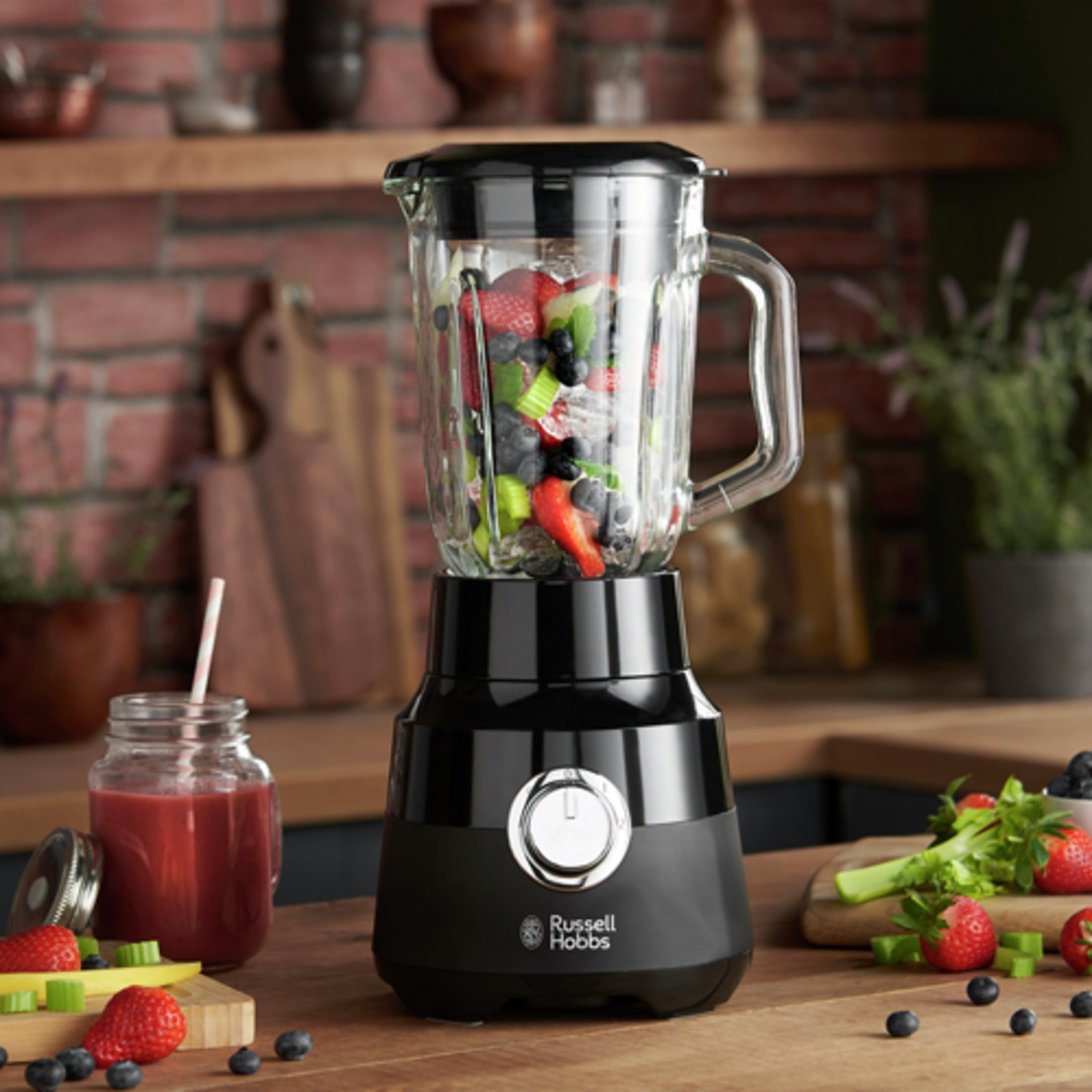 Ein schwarzer Mixer mit Früchten und ein Glas Saft stehen auf einem Holztisch.