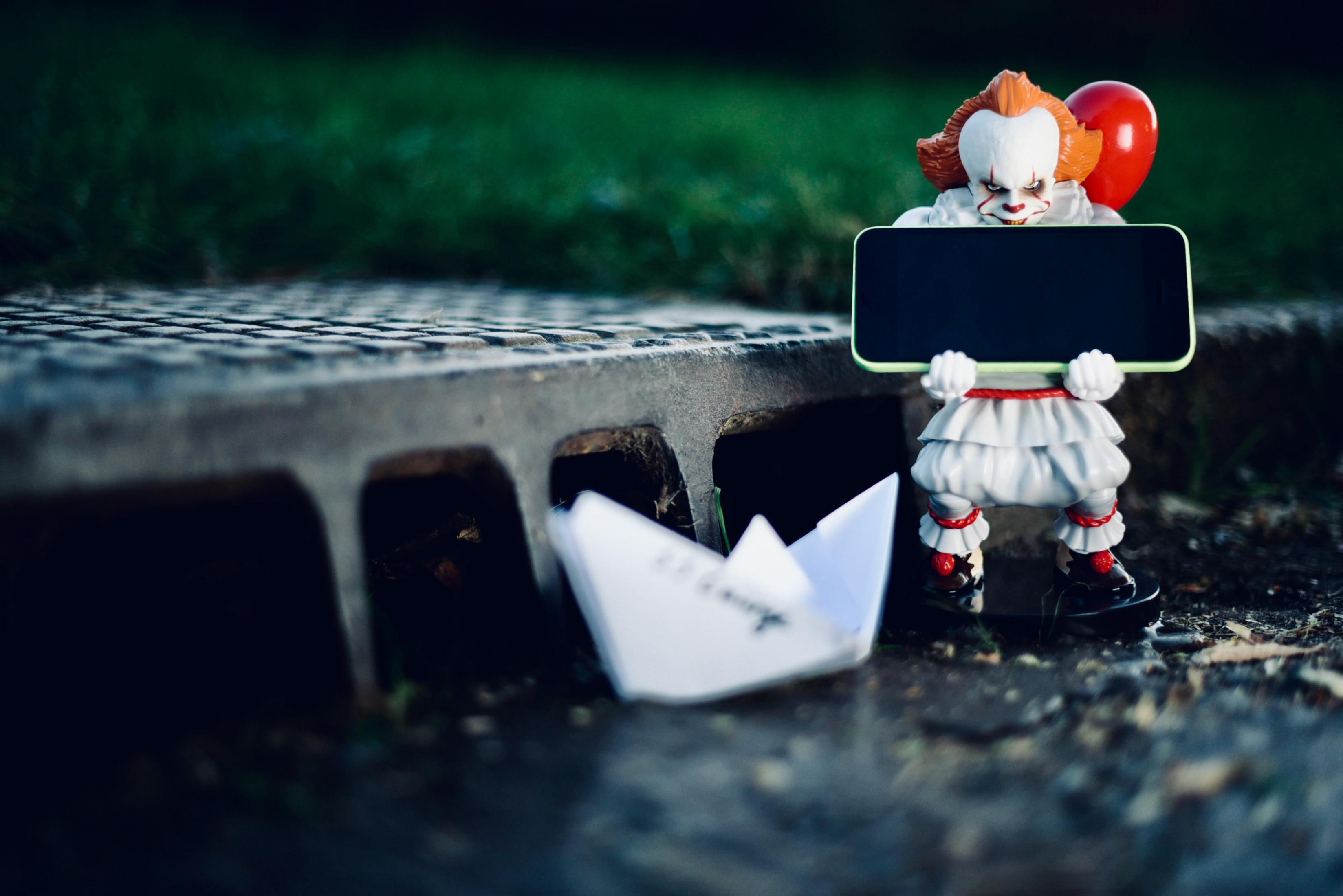 Eine Pennywise-Figur mit rotem Ballon hält ein Smartphone vor einem Gully.