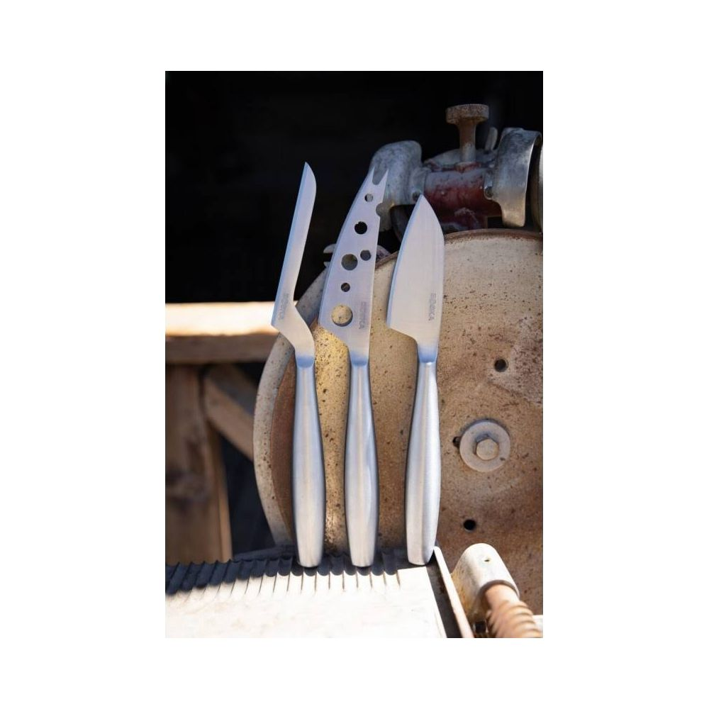 Three silver cheese knives with polished handles stand before a rusty metal object.