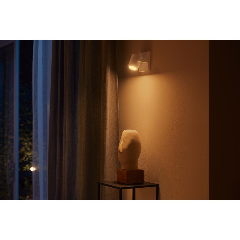 Kremowa lampa ścienna oświetla nowoczesny stół z białą abstrakcyjną rzeźbą, obok ciemnej zasłony.