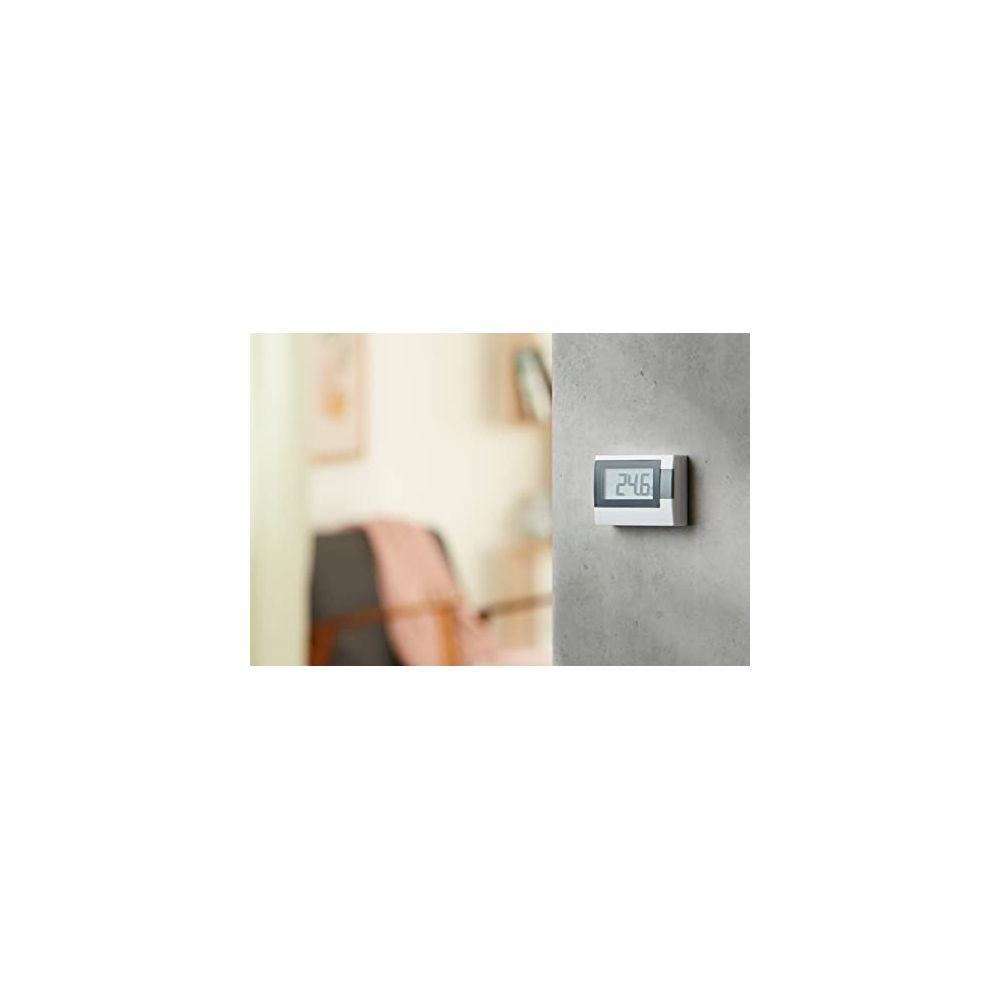 Ein weißer Thermostat mit digitaler Anzeige (24.6) an einer grauen Wand, mit unscharfem Wohnzimmer im Hintergrund.