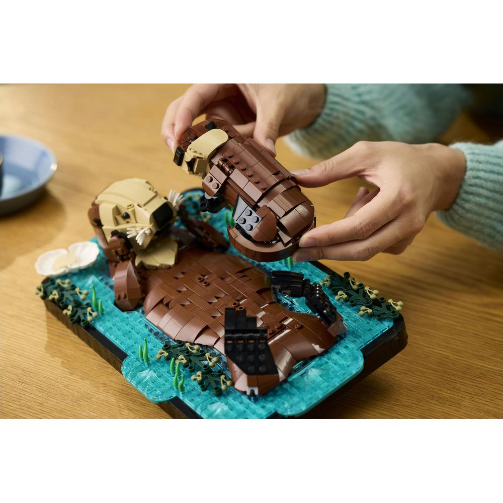 Ręce składają brązową wydrę Lego na niebieskiej podstawie wodnej z zielonymi roślinami.