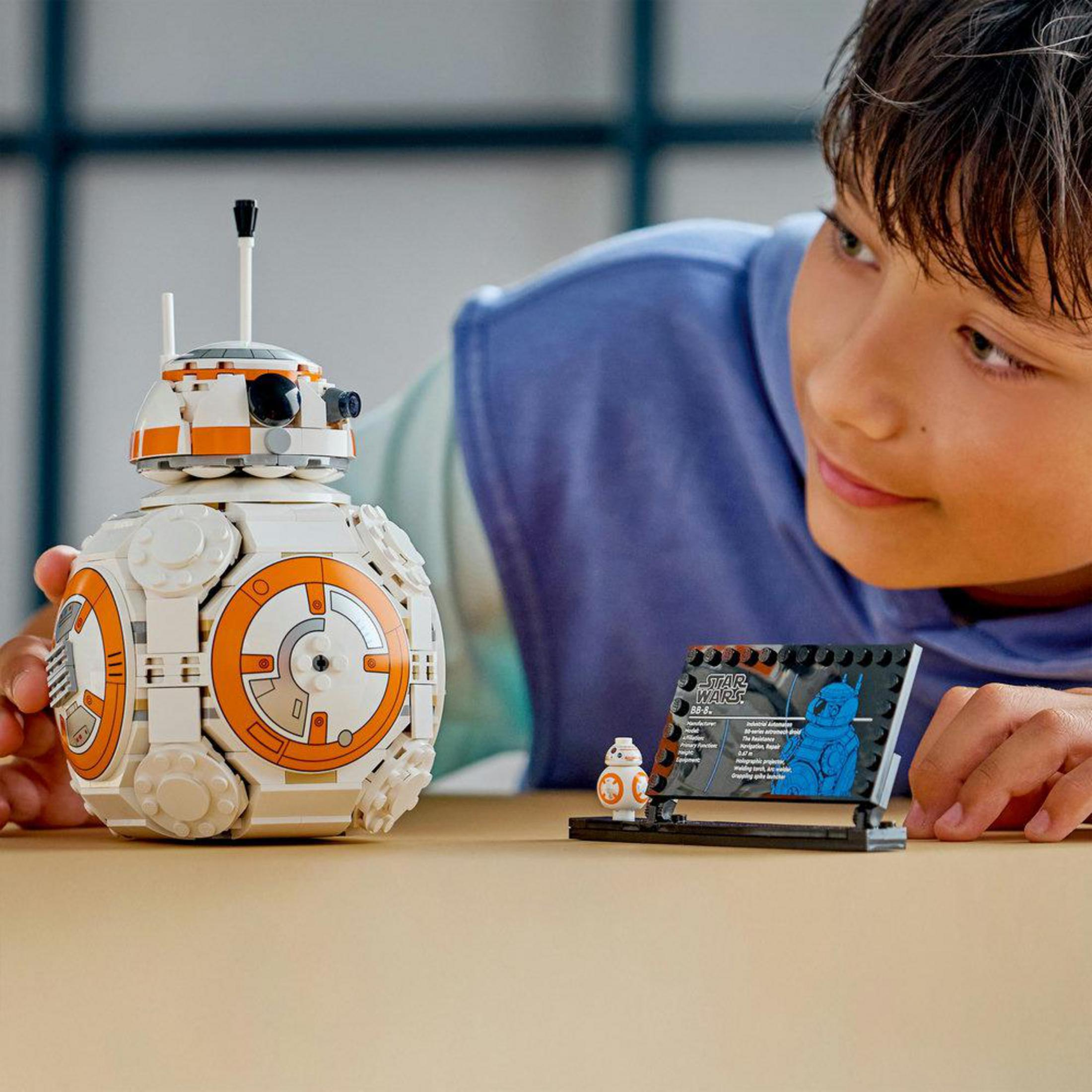 Chłopiec w niebieskiej bluzie patrzy na duży i mały model Lego BB-8 na stole.