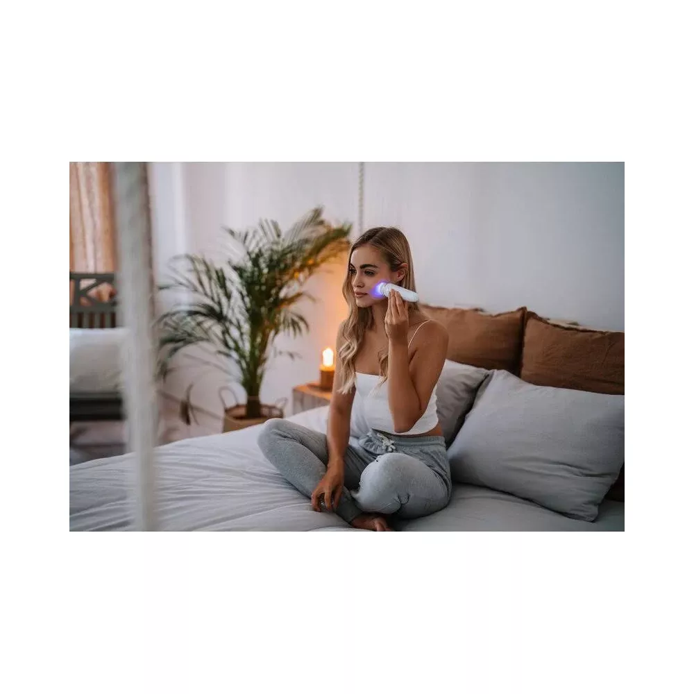 Eine Frau sitzt auf einem Bett und benutzt ein Hautpflegegerät mit blauem Licht.
