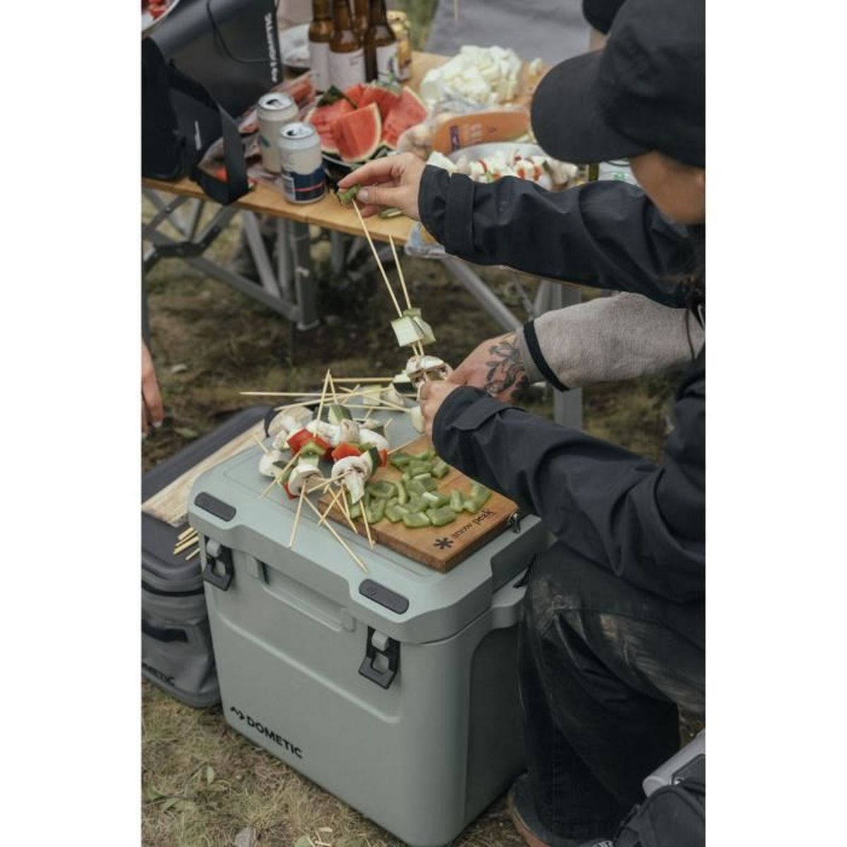 Persone preparano spiedini con verdure e funghi sul coperchio di un frigorifero portatile.