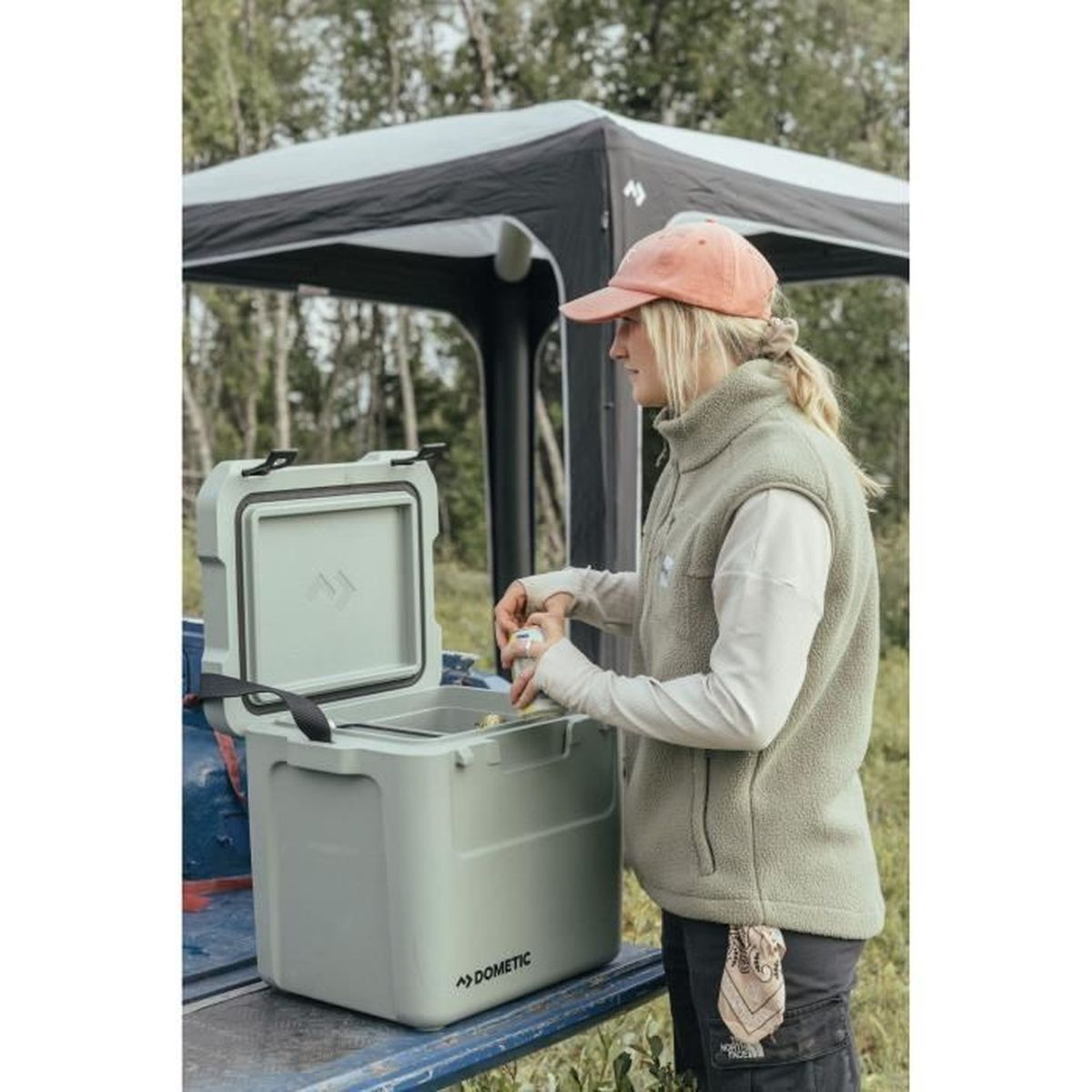 Una donna con cappello e gilet apre un box frigo vicino a una tenda.