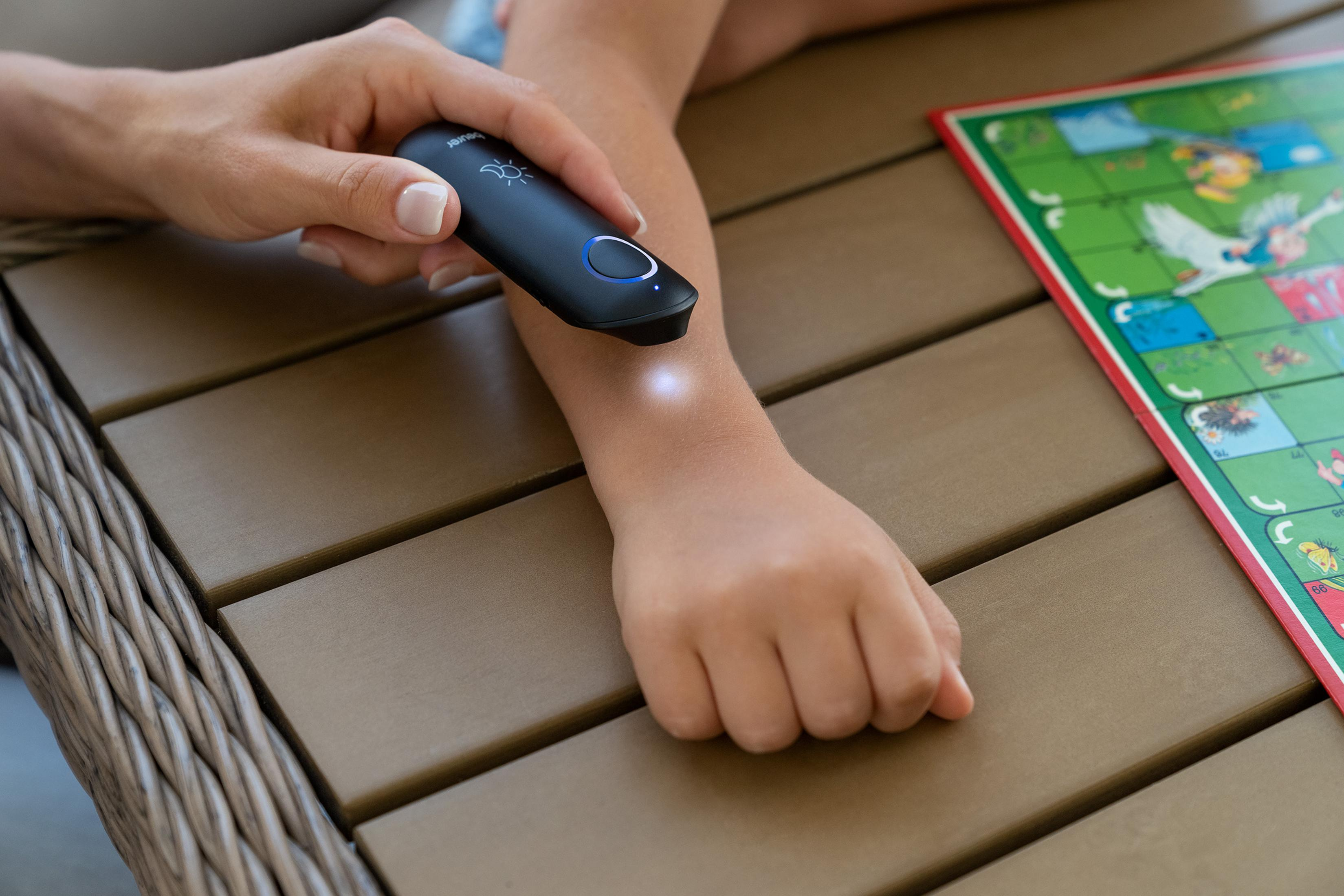 Eine Hand hält ein schwarzes Gerät über den Arm eines Kindes, ein Spielbrett liegt daneben.