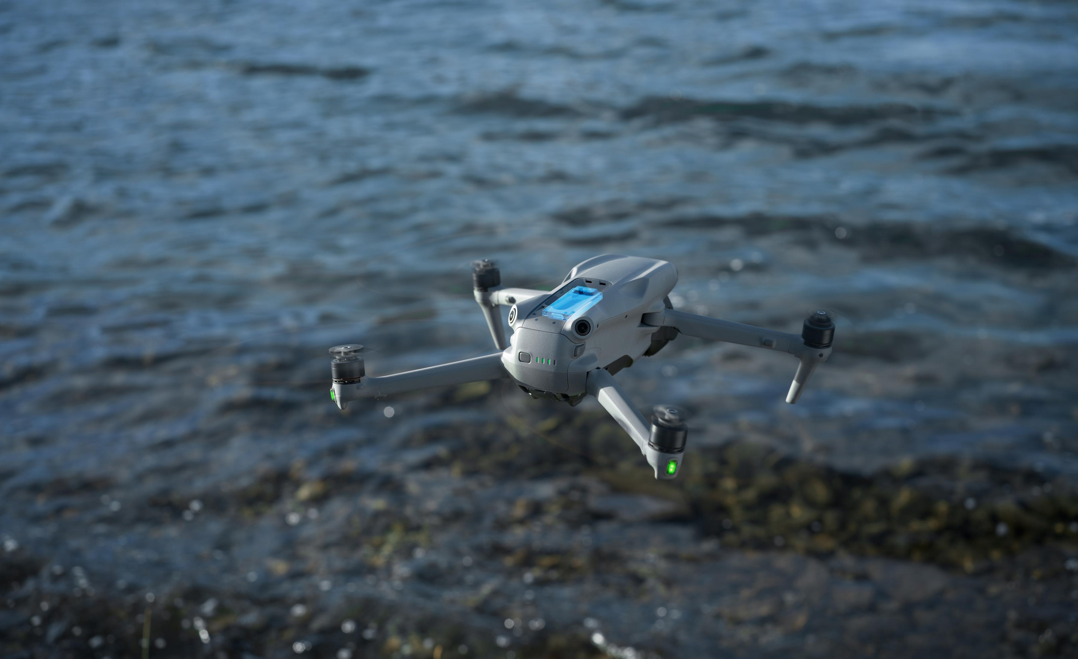 Szary dron leci nad wodą z kamieniami na brzegu.