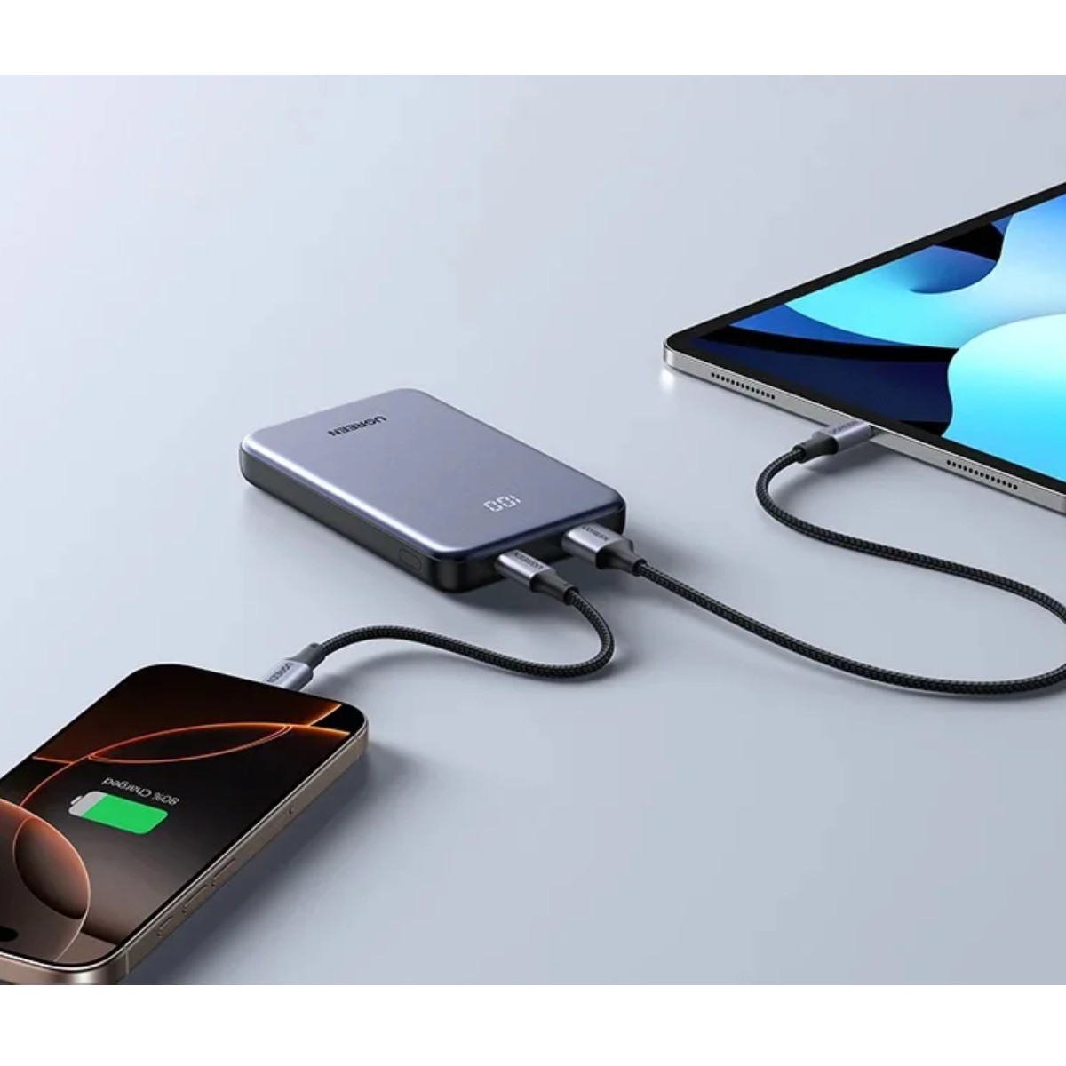 Szara powerbank ładuje iPhone'a i tablet za pomocą kabli.