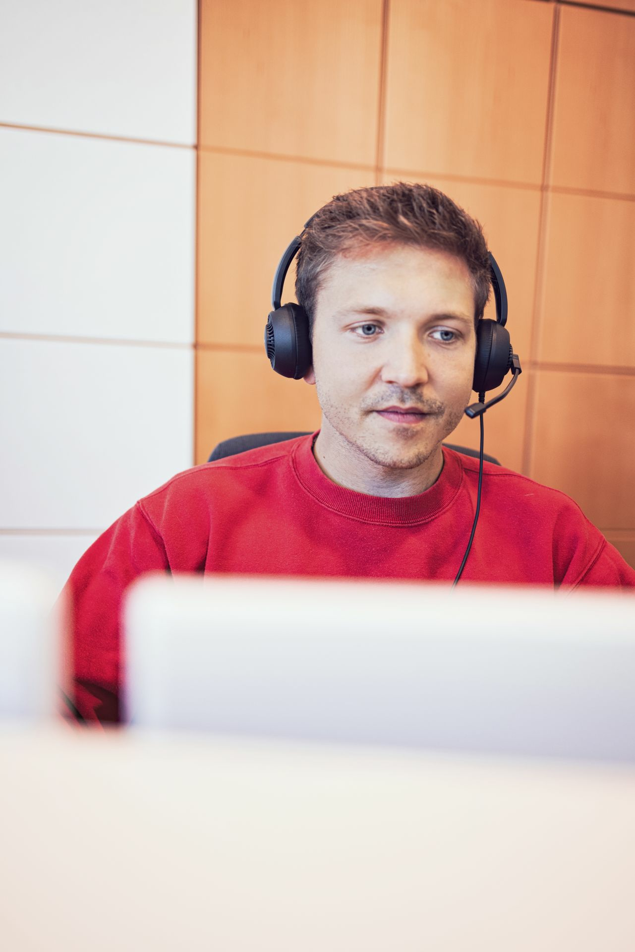 Man wearing headphones and a red sweater looks at a computer screen.