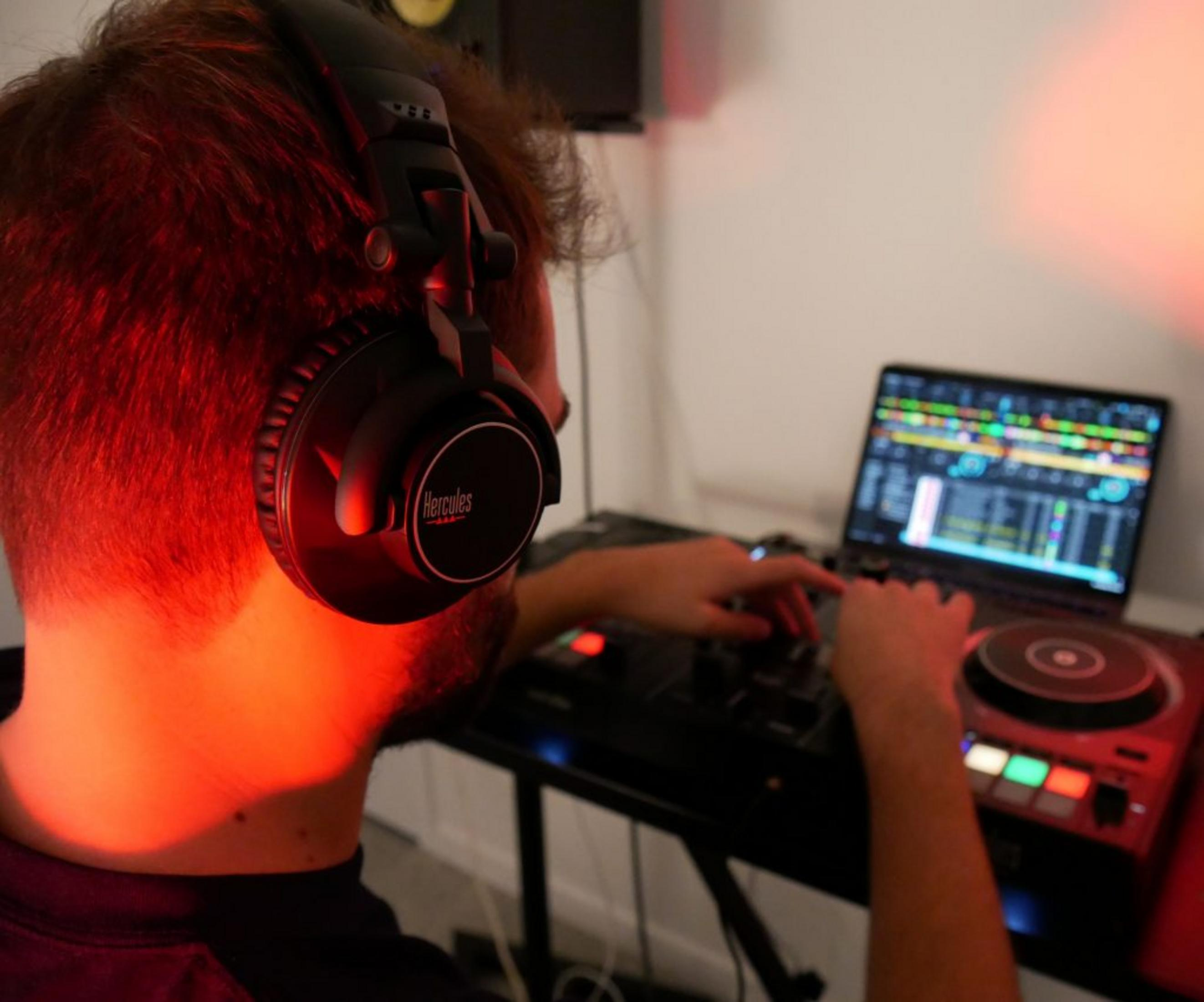 DJ mit Kopfhörern mischt Musik auf einem Laptop und Controller, beleuchtet von rotem Licht.