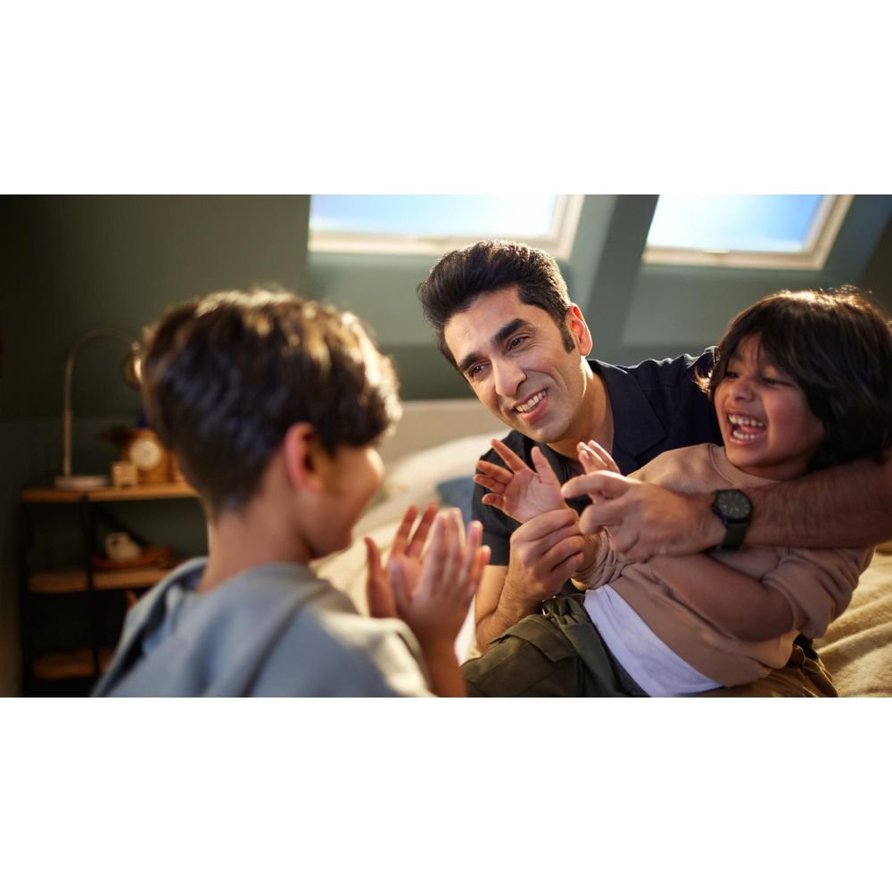 Mężczyzna bawi się z dwójką śmiejących się dzieci na łóżku. Niebo widoczne przez okna dachowe.