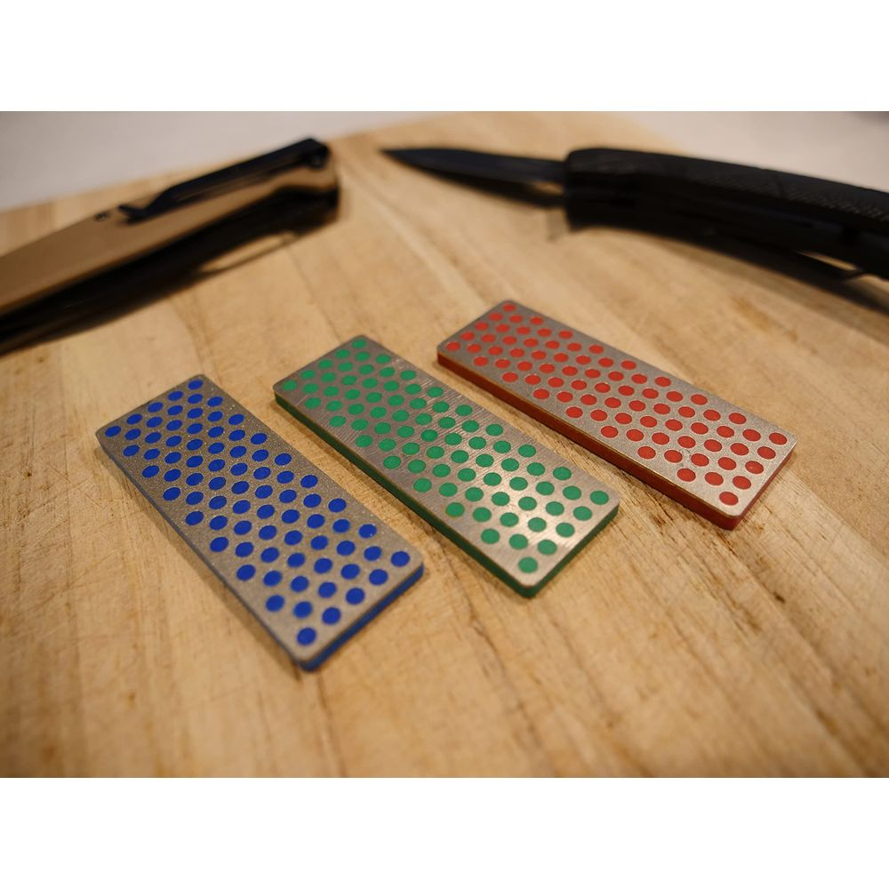 Trois pierres à aiguiser, bleue, verte et rouge, sont posées sur une planche en bois près d'un couteau.