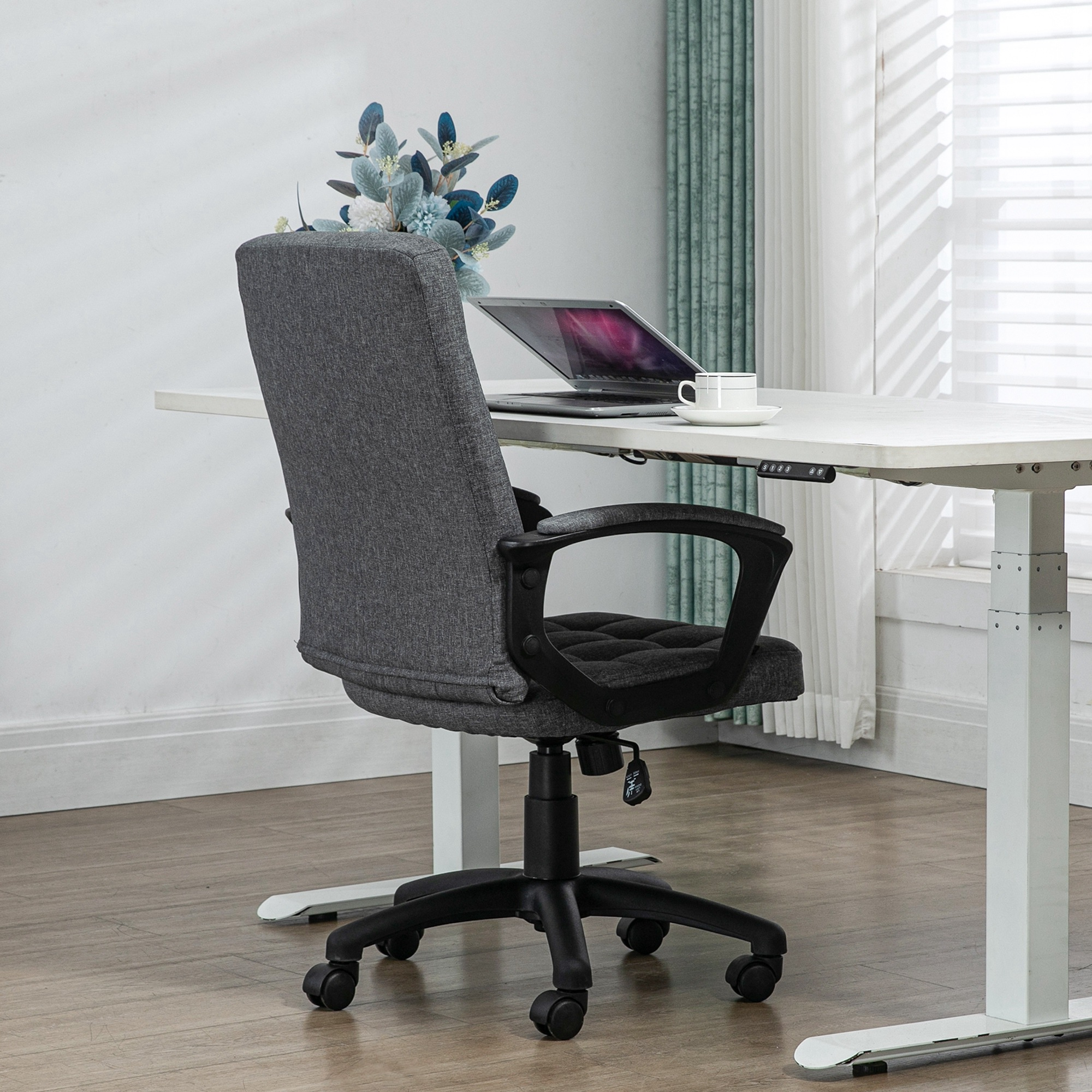 Una silla de oficina gris junto a un escritorio elevable con portátil y taza de café.