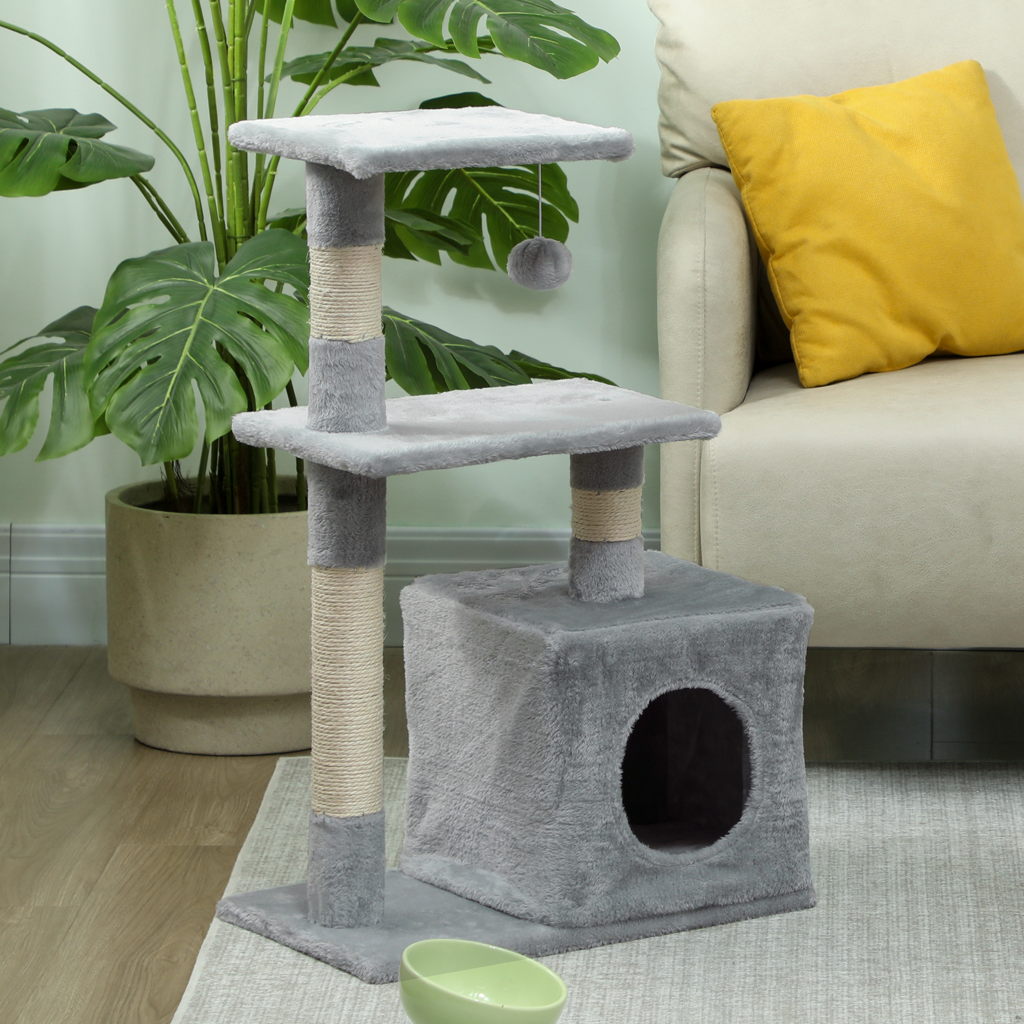 Árbol rascador gris con juguete y casa, junto a planta y sofá.