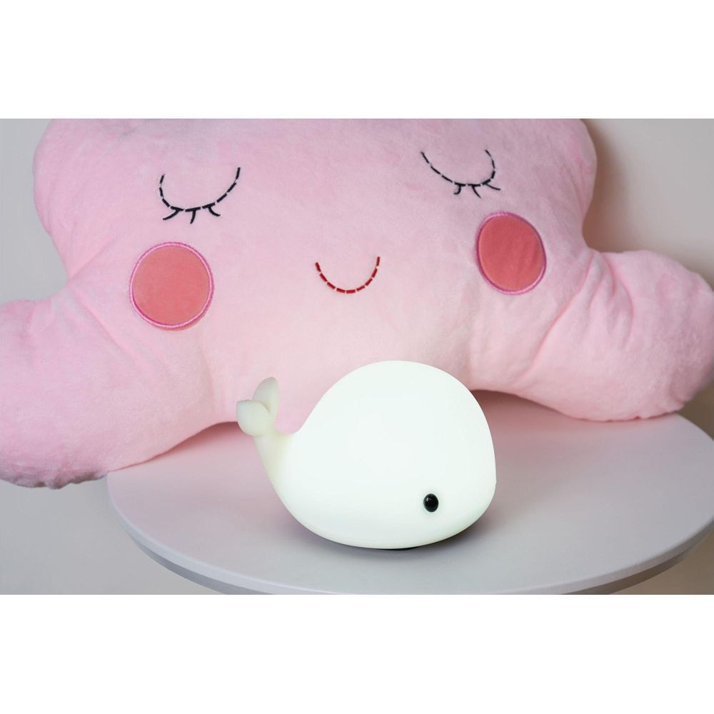 Mała biała lampka nocna w kształcie wieloryba przed dużą różową poduszką.