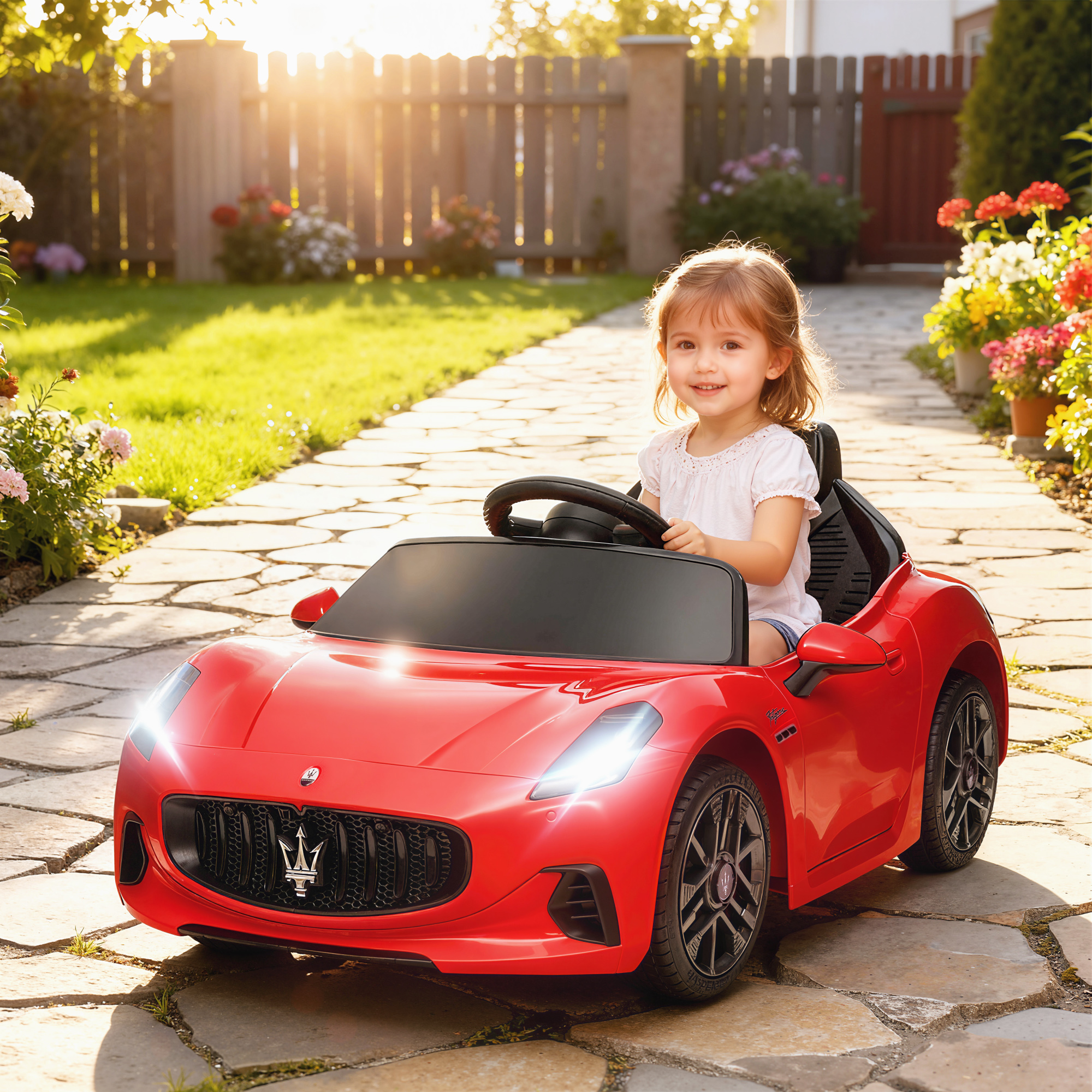 Una niña sonriente con camisa blanca conduce un coche de juguete rojo por un camino de piedra en un jardín.