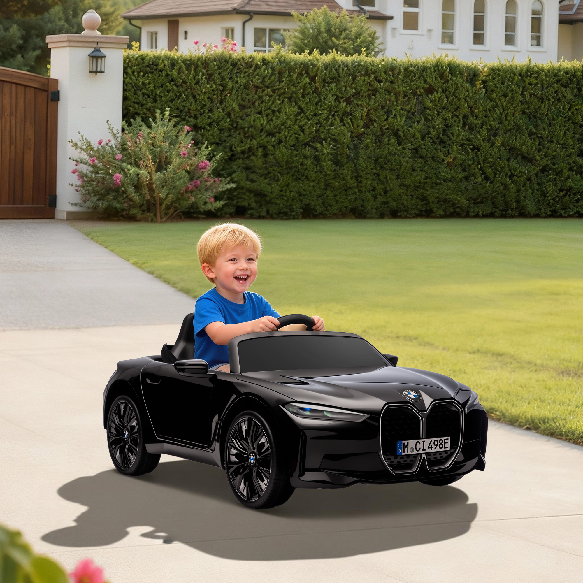Un niño sonriente conduce un coche de juguete BMW negro en un camino de entrada frente a una casa.