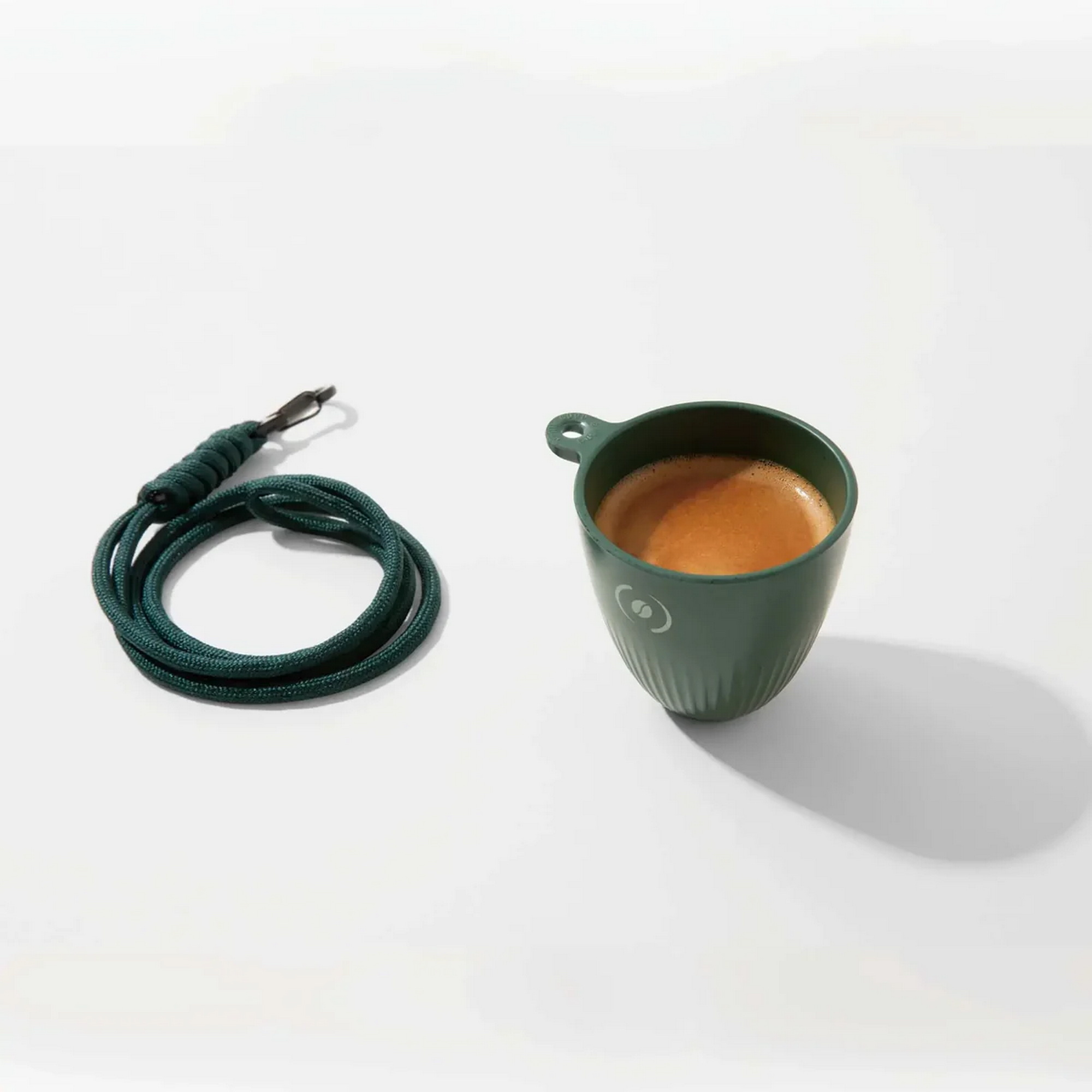 Ein grüner Becher mit Espresso neben einer grünen Kordel auf weißem Grund.