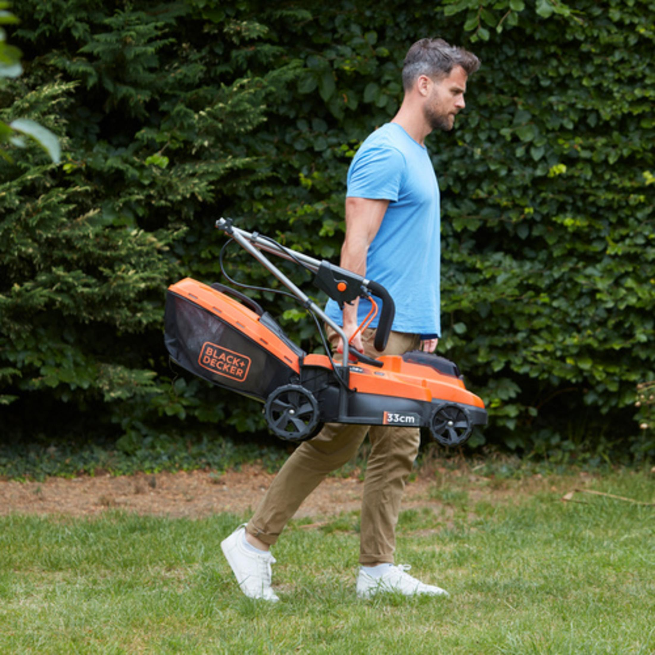Mann trägt einen orangefarbenen Rasenmäher durch einen Garten mit grünem Laub.