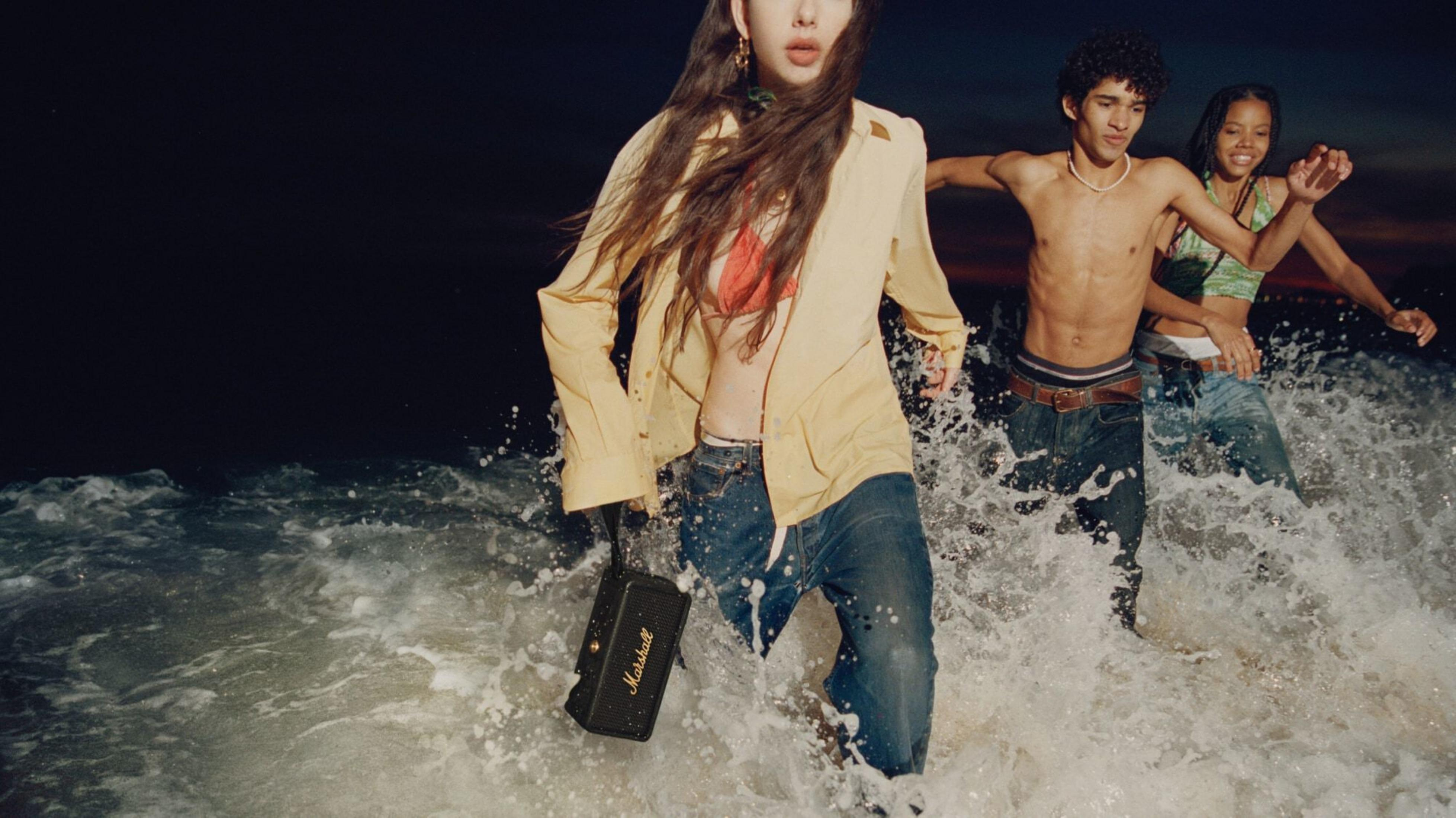 Drei junge Leute laufen im Meer mit Wellen und Gischt, eine Person trägt einen Lautsprecher.