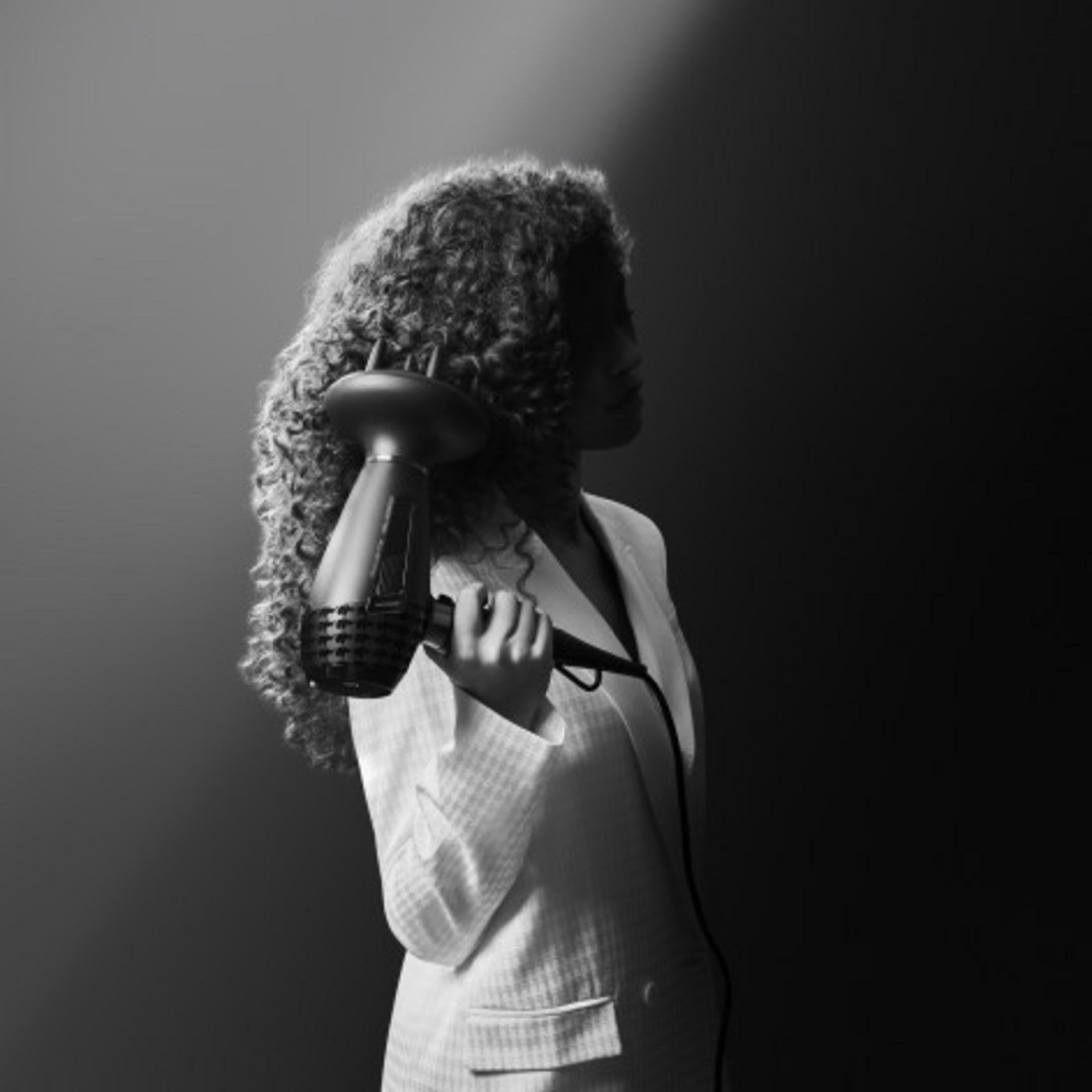 Kobieta z kręconymi włosami trzyma suszarkę z dyfuzorem w jasnej marynarce.