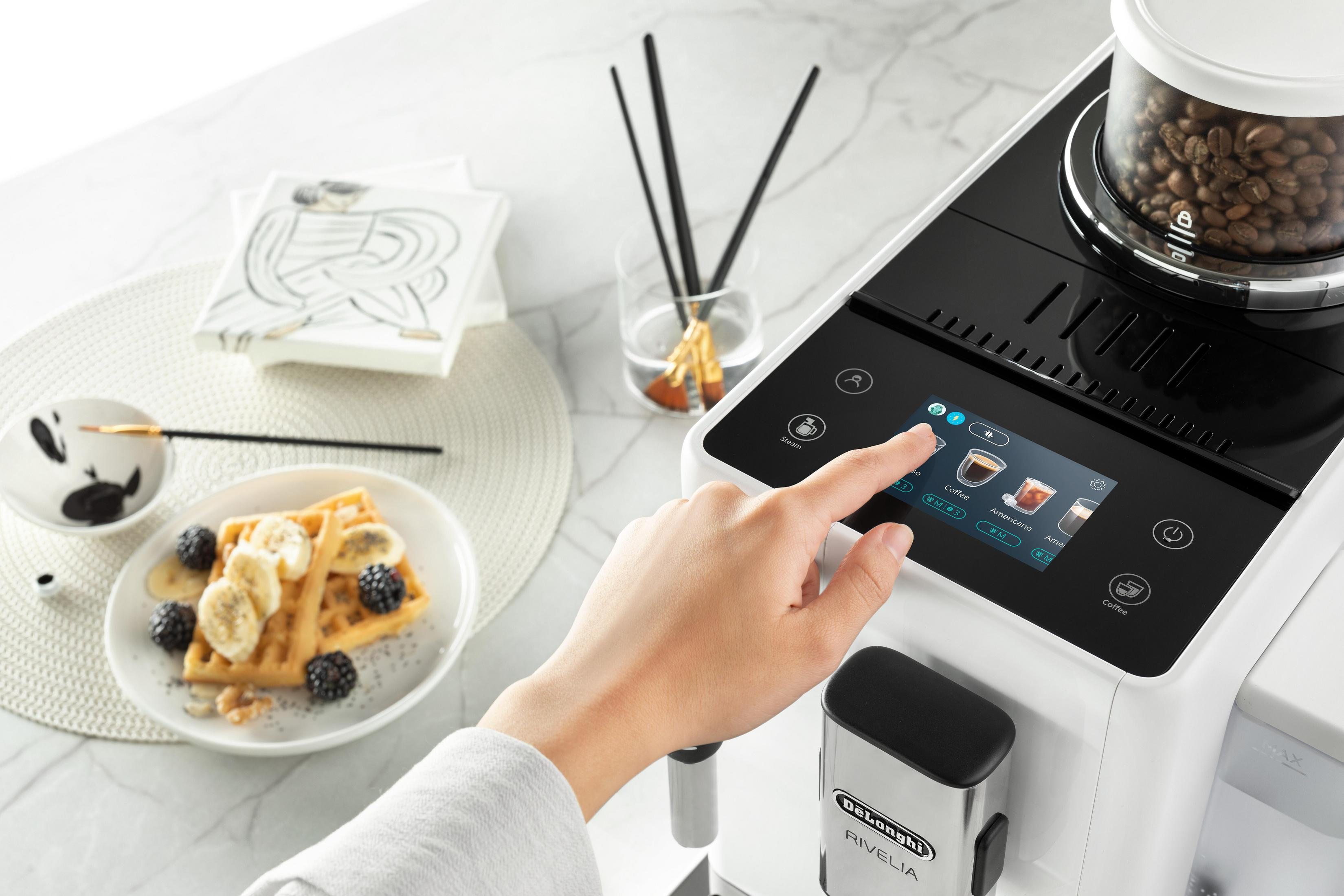 Dłoń obsługuje biały ekspres do kawy z ekranem dotykowym. Obok danie z goframi.