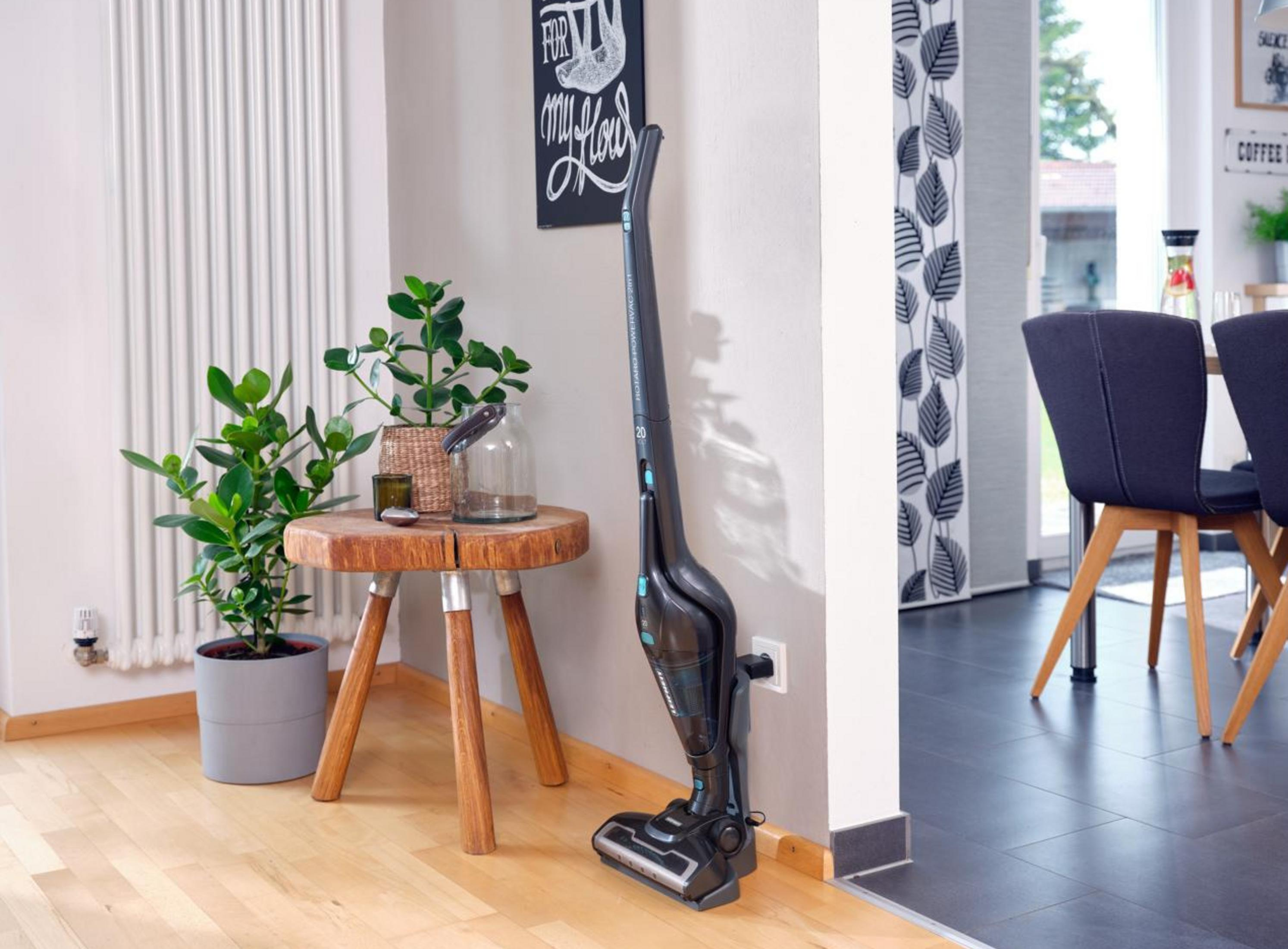 Ciemny odkurzacz bezprzewodowy ładuje się przy ścianie obok małego stolika z rośliną.