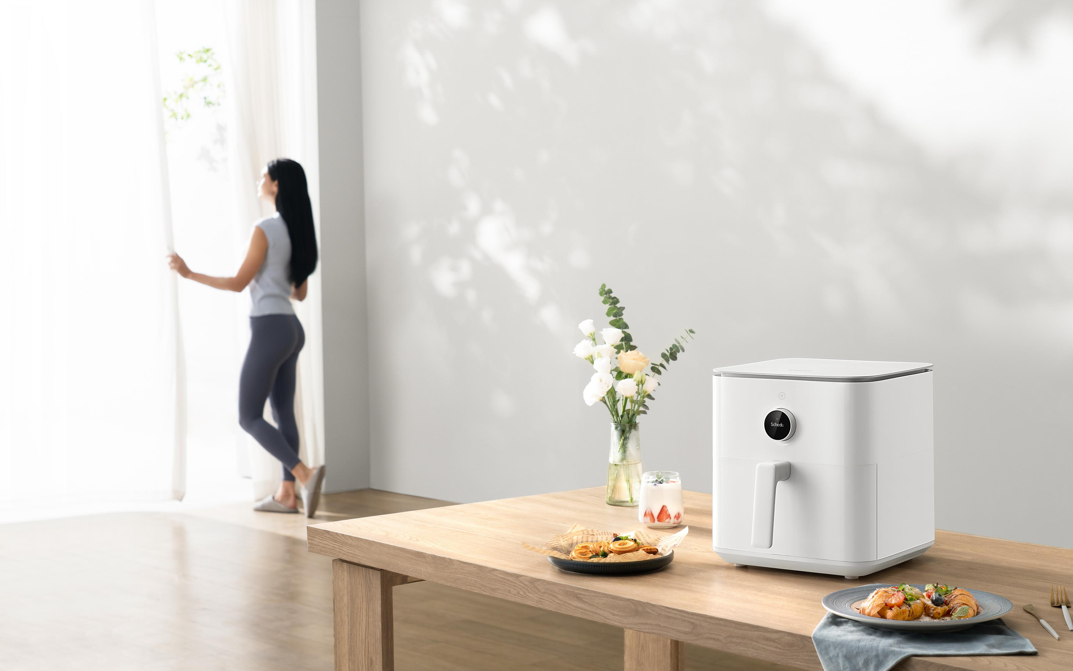 Kobieta otwiera zasłony. Na stole biała frytkownica beztłuszczowa, kwiaty i jedzenie.