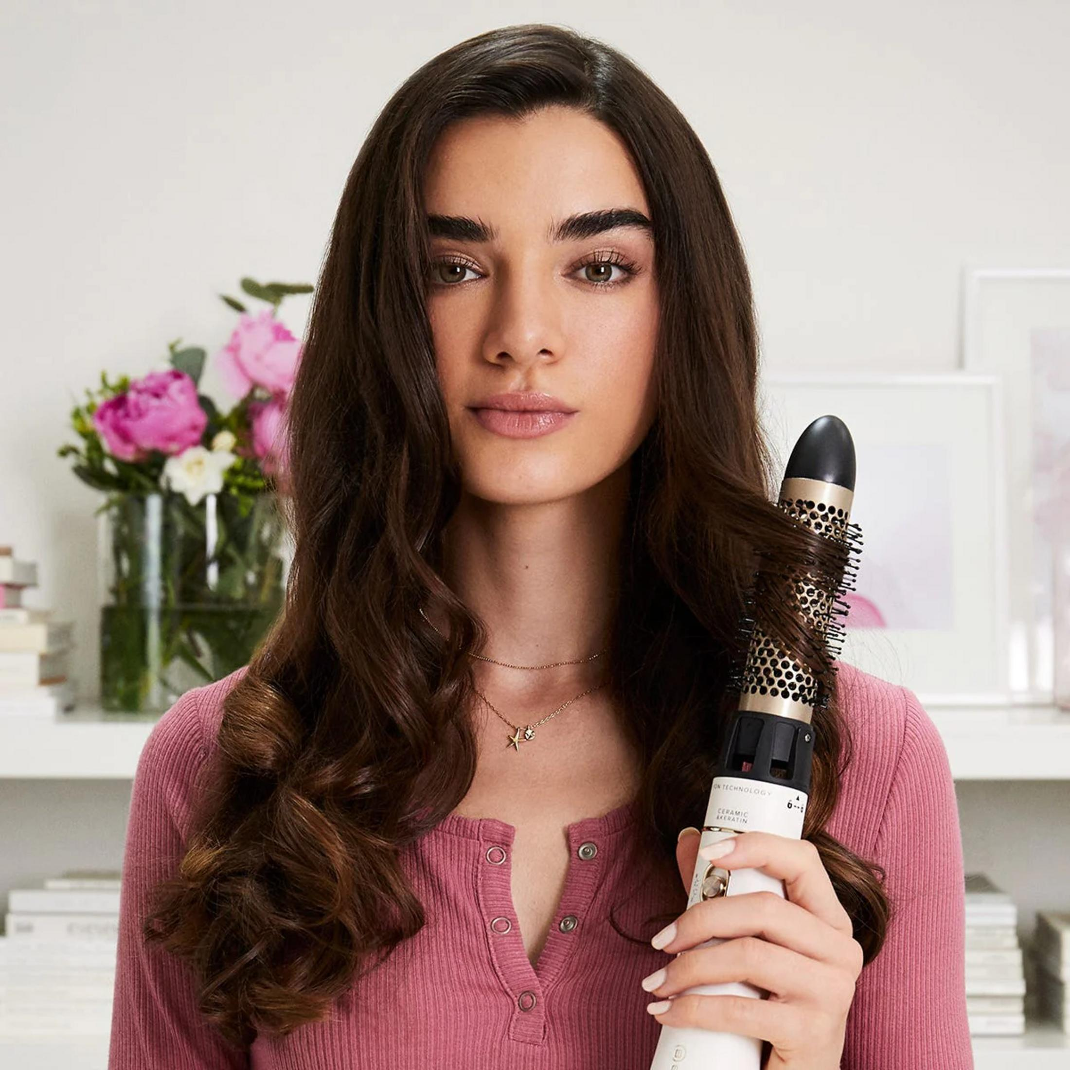 Kobieta z brązowymi włosami trzyma lokówkę. W tle kwiaty.