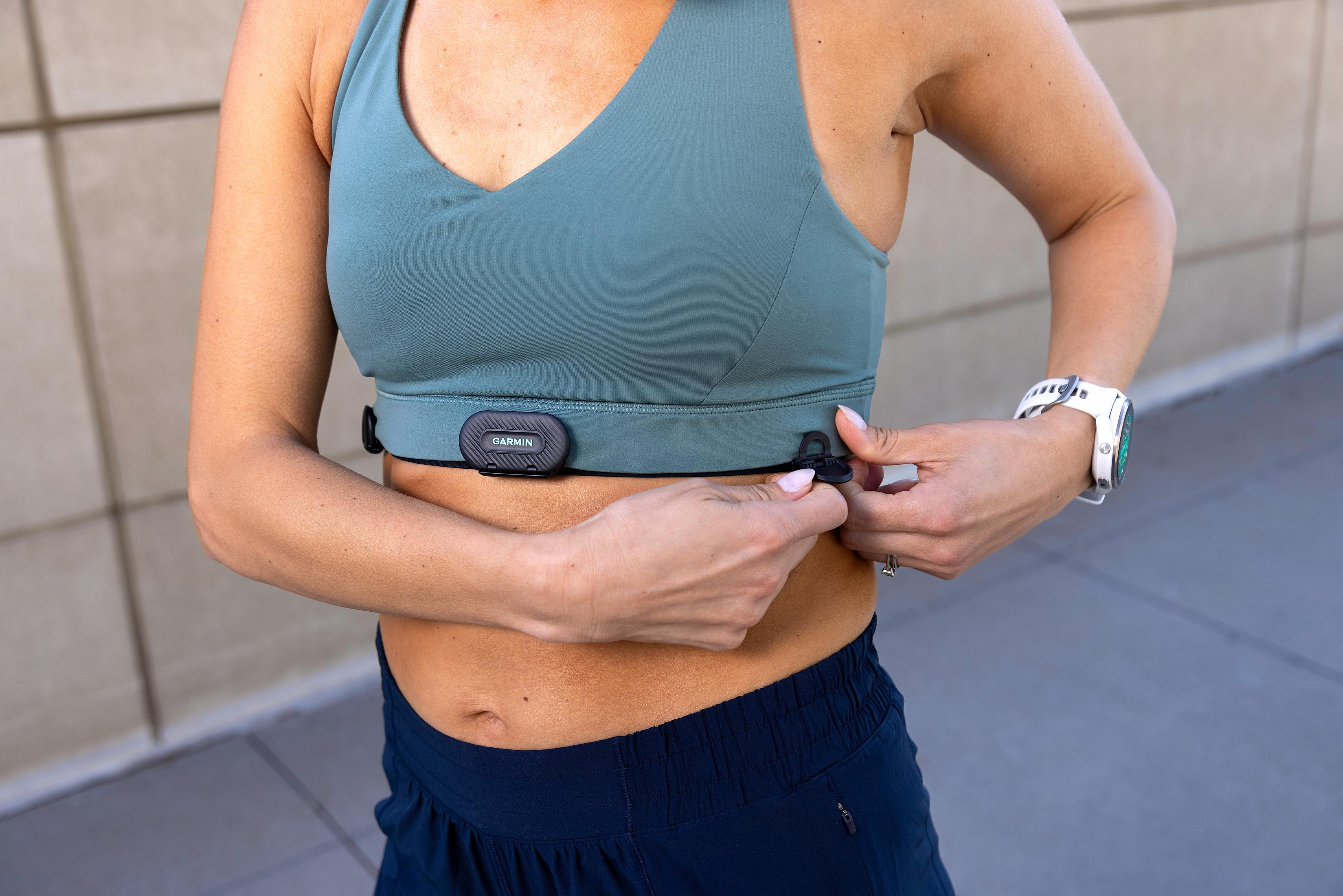 Bir kadın Garmin kalp atış hızı monitörü bandını takıyor. Spor sütyeni ve mavi şort giyiyor.