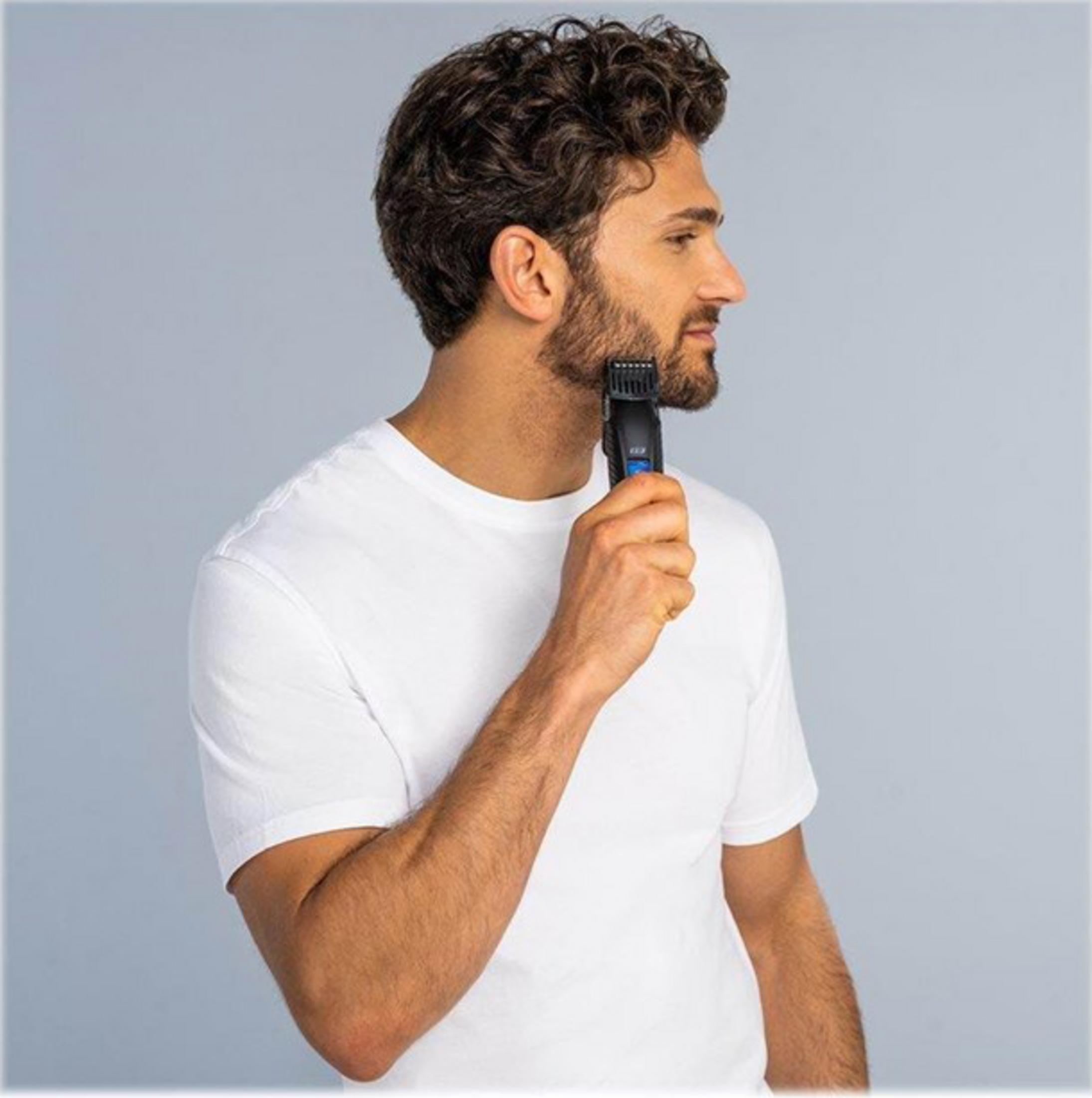 Hombre en camiseta blanca recortando barba con recortadora eléctrica negra sobre fondo gris.