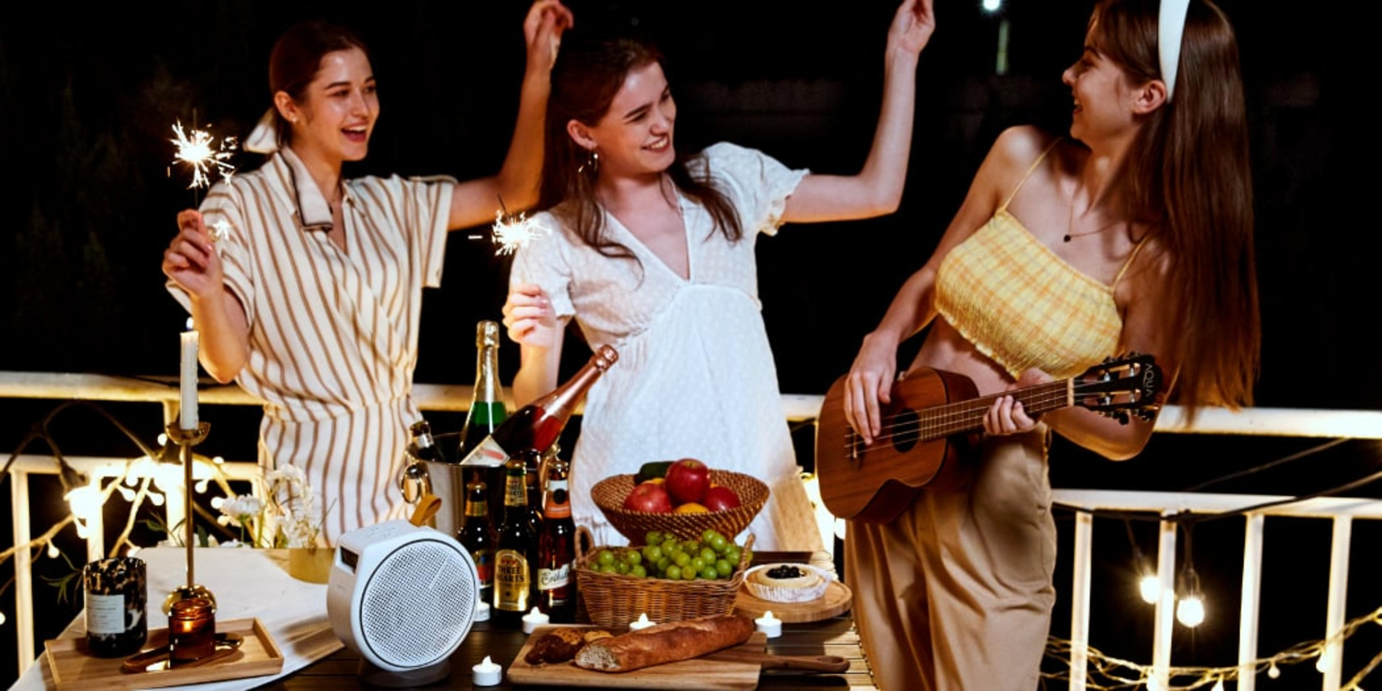 Drei Frauen feiern mit Wunderkerzen und Ukulele auf einem Balkon bei Nacht.