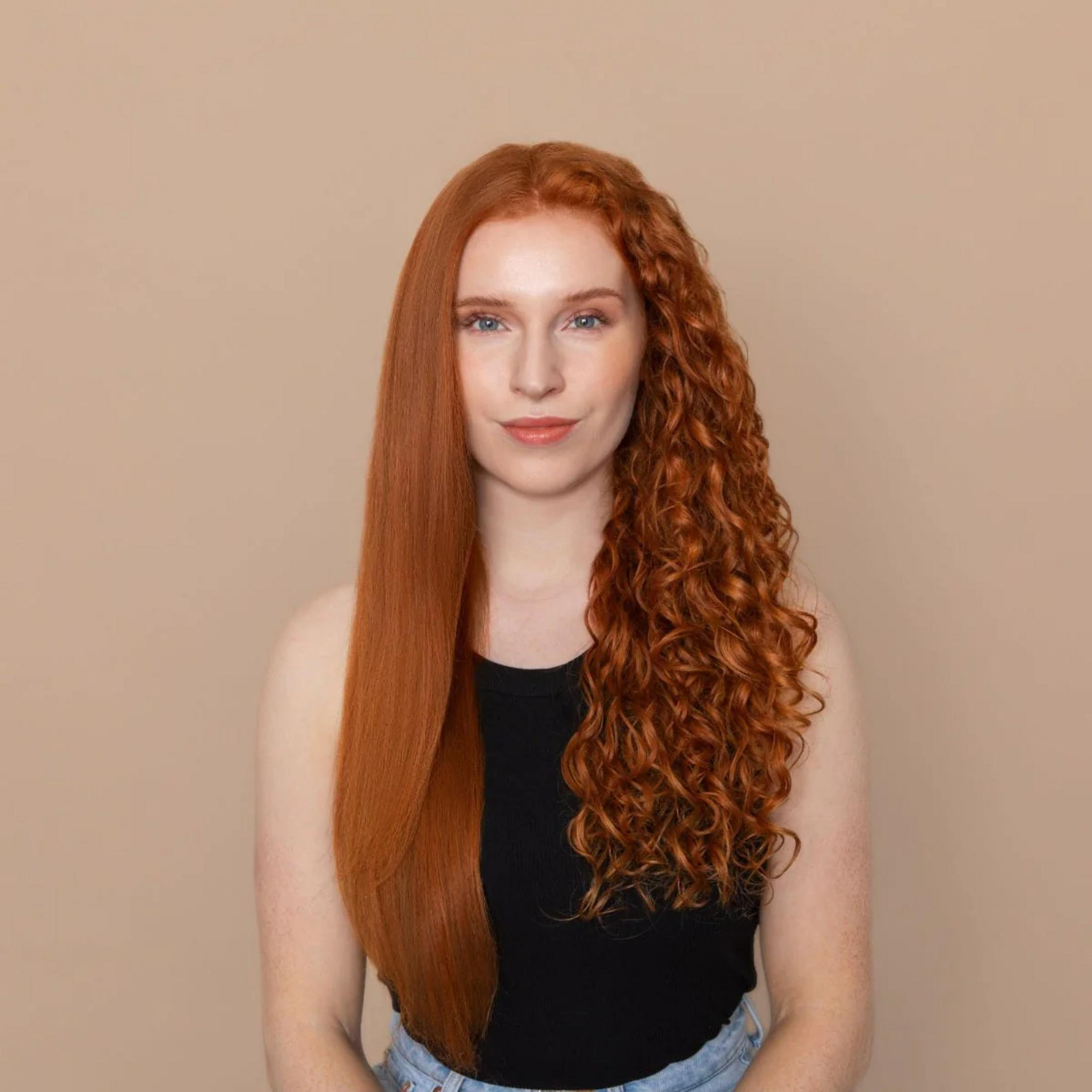 Donna con capelli rossi, un lato liscio, l'altro riccio, su sfondo beige.