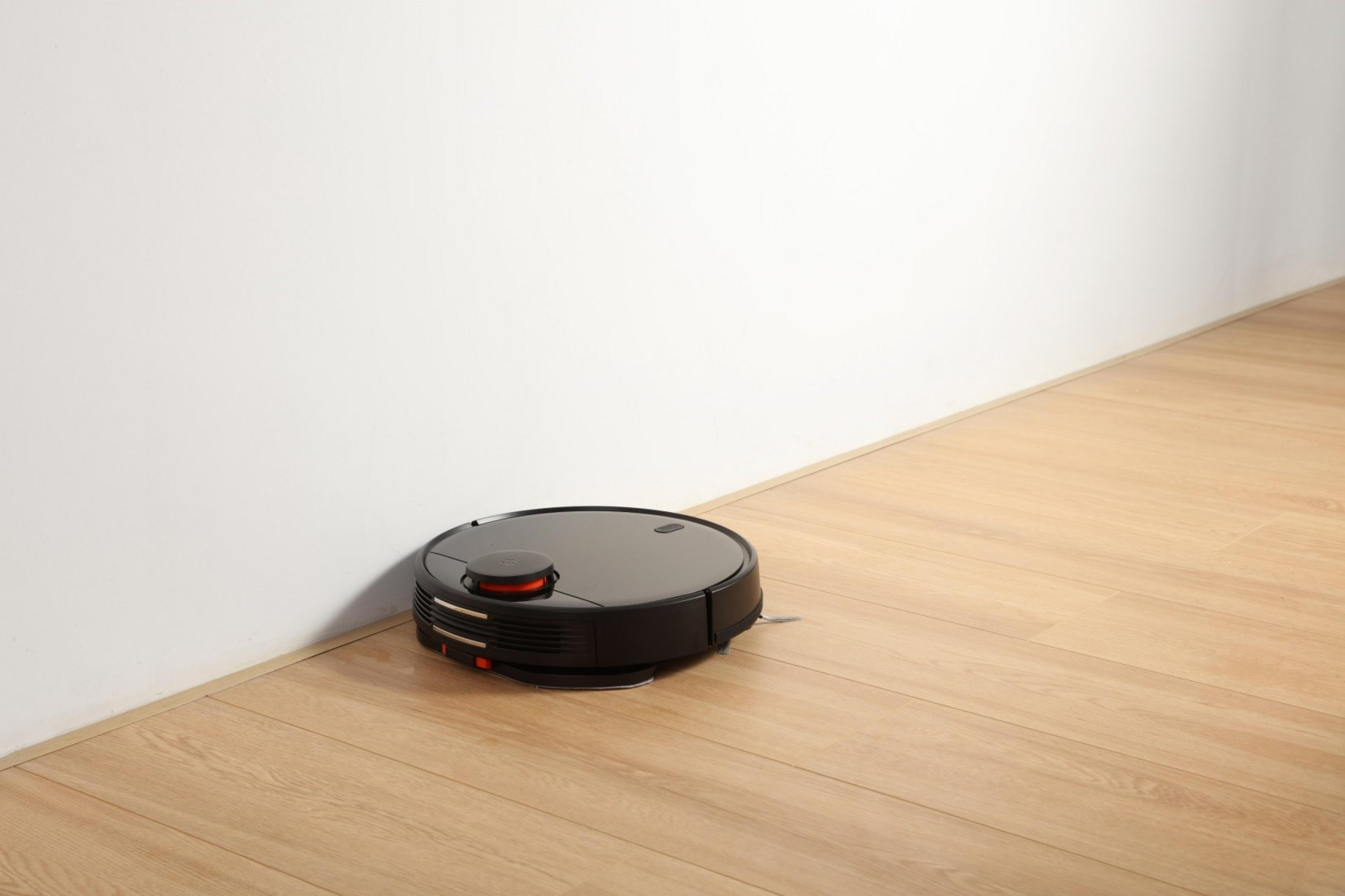 Un robot aspirador negro limpia un suelo de madera junto a una pared blanca.